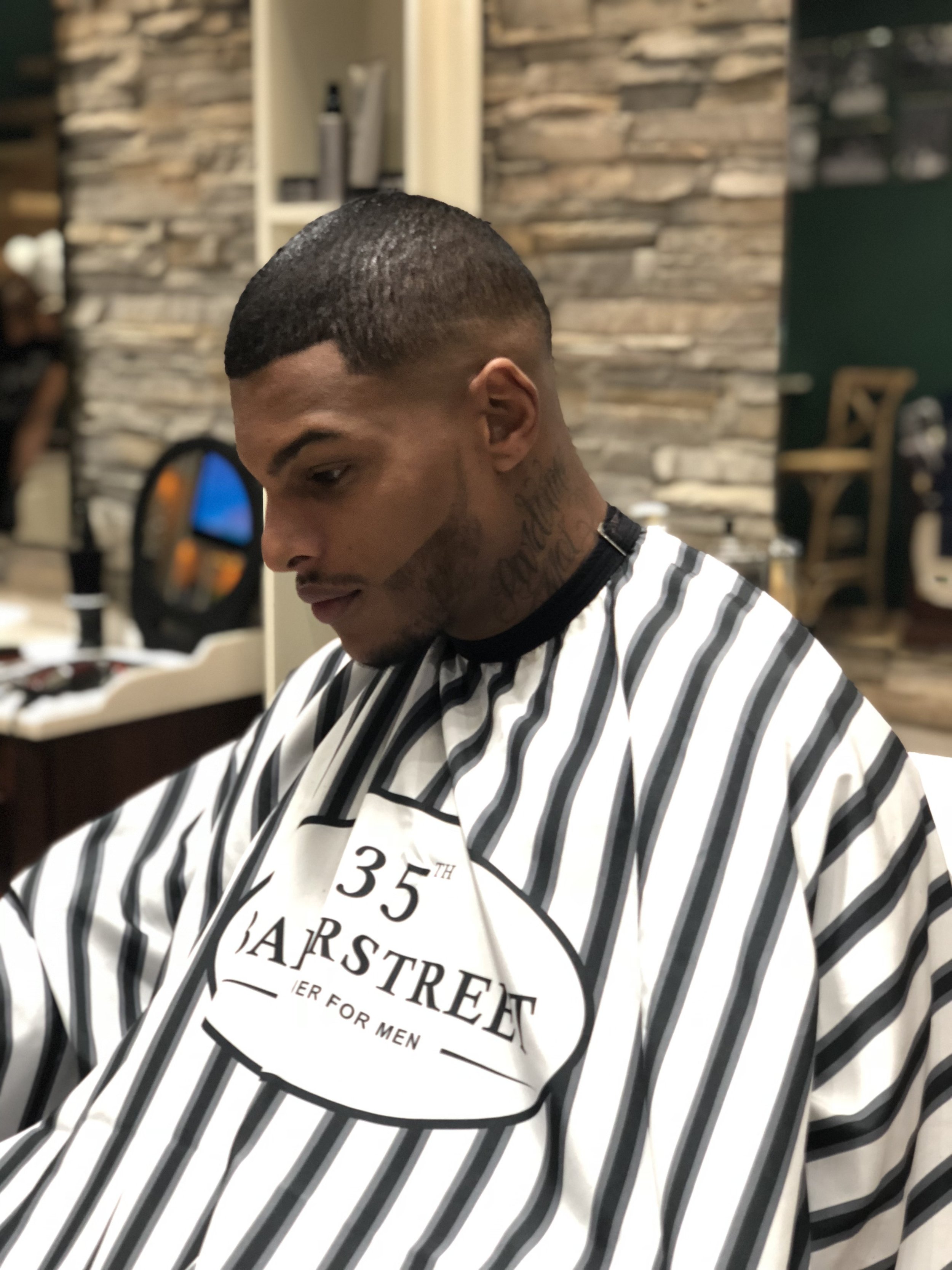 Un homme assis dans un salon de coiffure, portant une cape de barbier rayée noir et blanc avec le texte '35th Street Barber'. Best barber shop salon de coiffure Marrakech