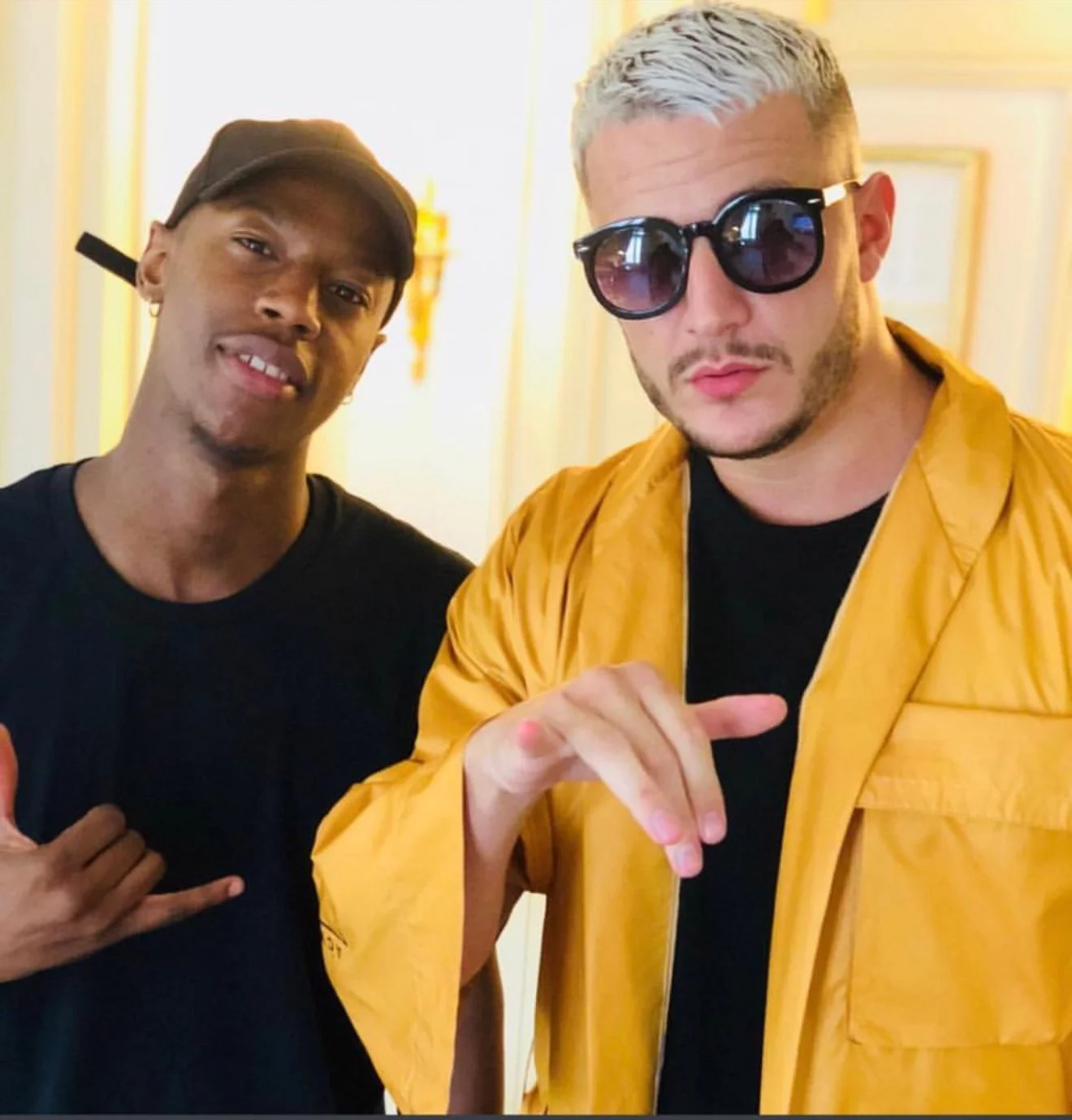 Deux hommes posant pour une photo, l'un porte un chapeau noir et un t-shirt noir, l'autre porte des lunettes de soleil noires et un manteau jaune, dans un intérieur éclairé. Best barber shop salon de coiffure Marrakech