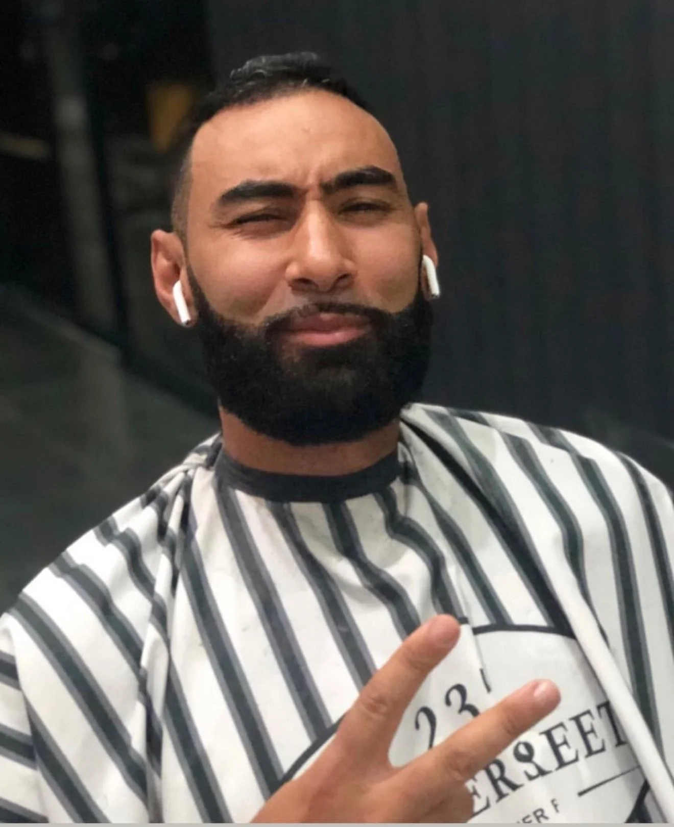 Un homme à la barbe, portant un équipement sans fil, fait un signe de paix avec deux doigts, portant une blouse de barbier à rayures. Best barber shop salon de coiffure Marrakech