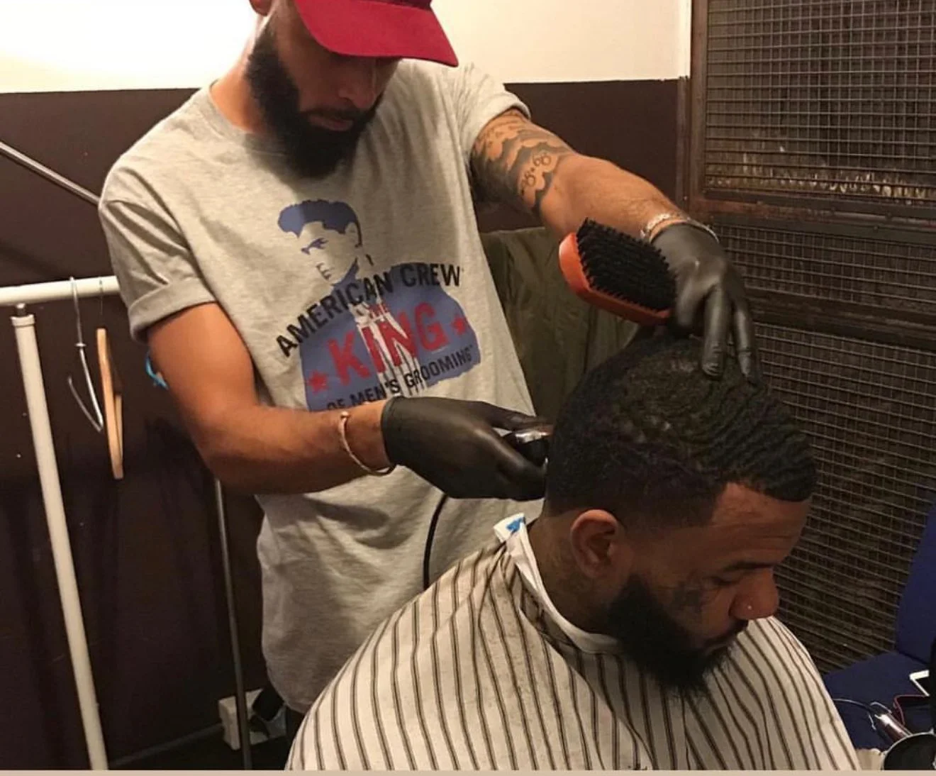 Un homme se fait couper les cheveux dans un salon de coiffure. Il porte une chemise rayée et regarde vers le bas. Le coiffeur, portant un t-shirt gris avec un logo et un chapeau rouge, utilise une tondeuse électrique pour couper la coiffure de l'homme, qui a les cheveux foncés et bouclés.