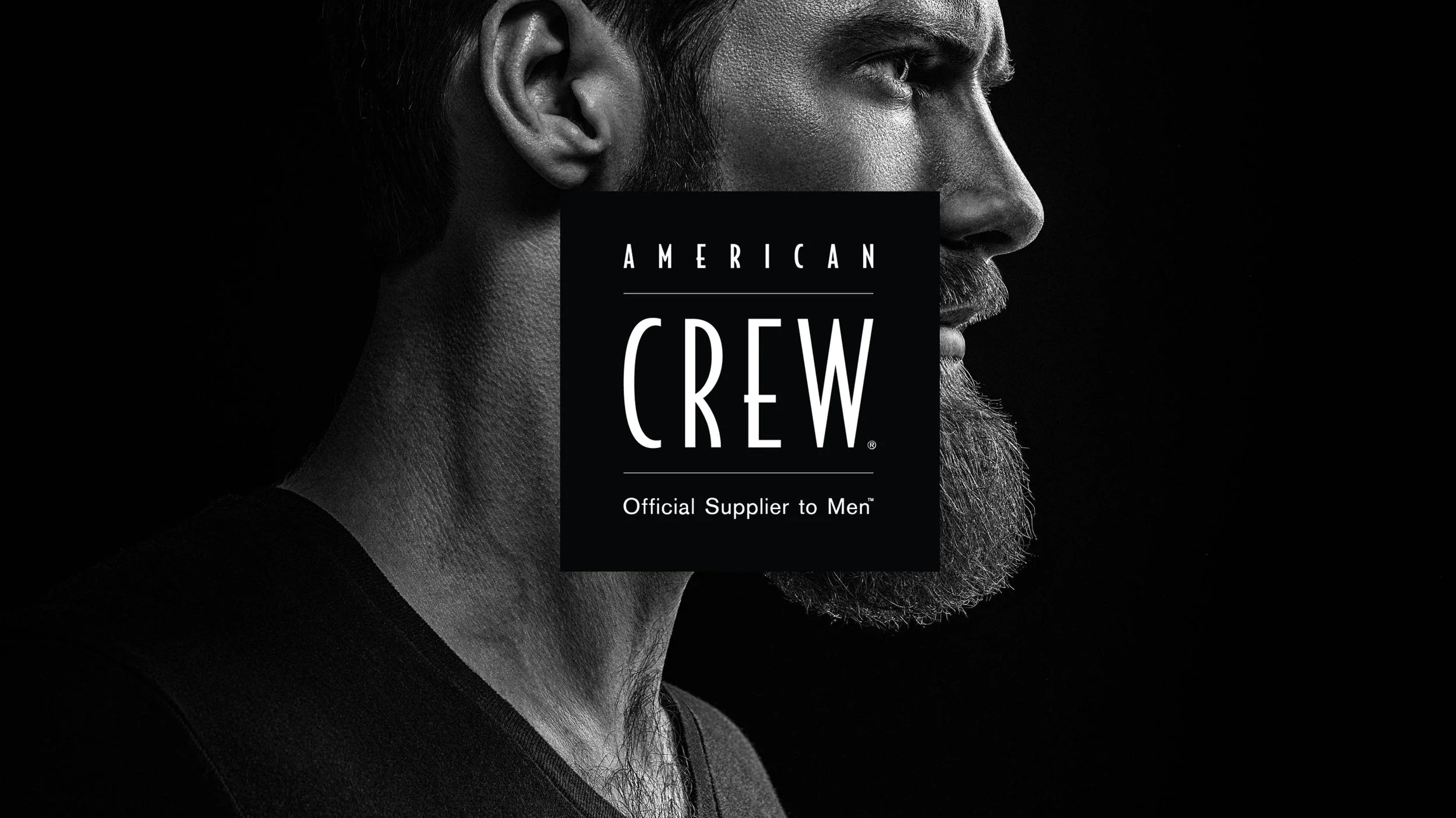 Un portrait en noir et blanc d'un homme avec une barbe et des cheveux courts, regardant vers la droite, avec un logo de American Crew au centre.