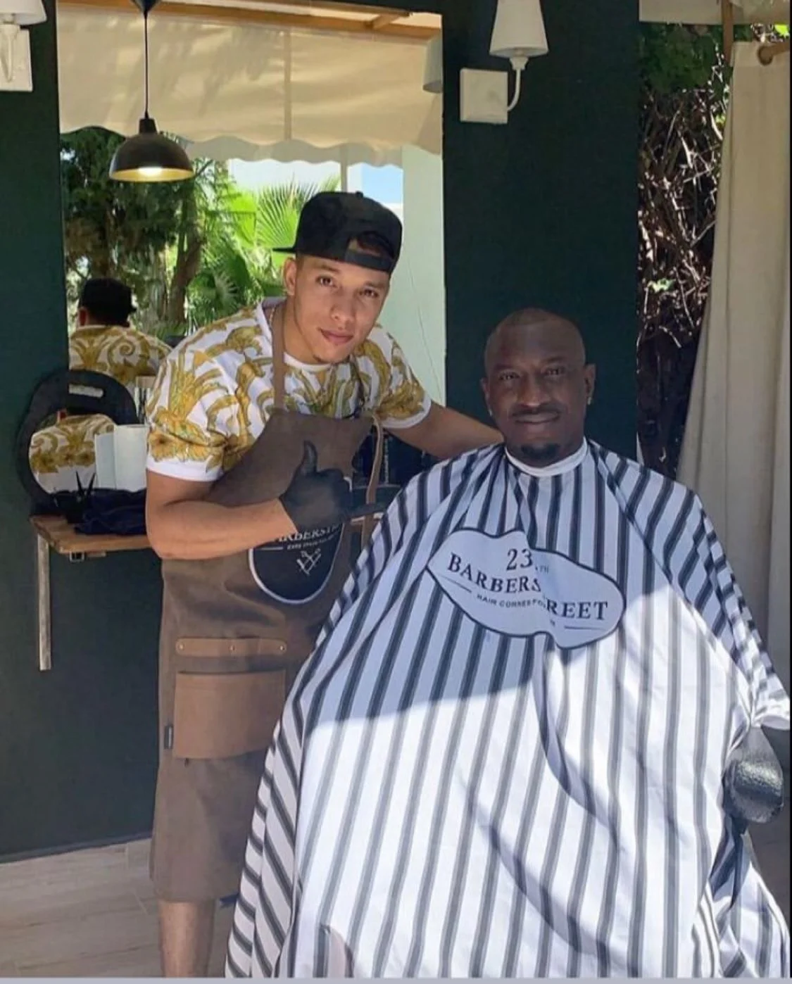 Un homme reçoit une coupe de cheveux dans un salon de coiffure, assis dans une cape de barber en noir et blanc. Un autre homme se tient derrière lui, portant un tablier d'apprenti coiffeur et un chapeau à rayures, dans un salon avec des plantes en arrière-plan.