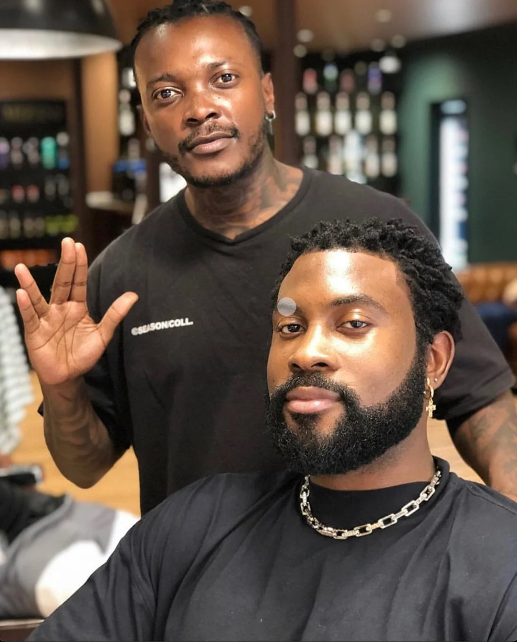 Deux hommes dans un salon de coiffure, l'un derrière avec une main levée, l'autre assis avec une barbe et des dreadlocks, portant une chaîne en argent. Best barber shop salon de coiffure Marrakech