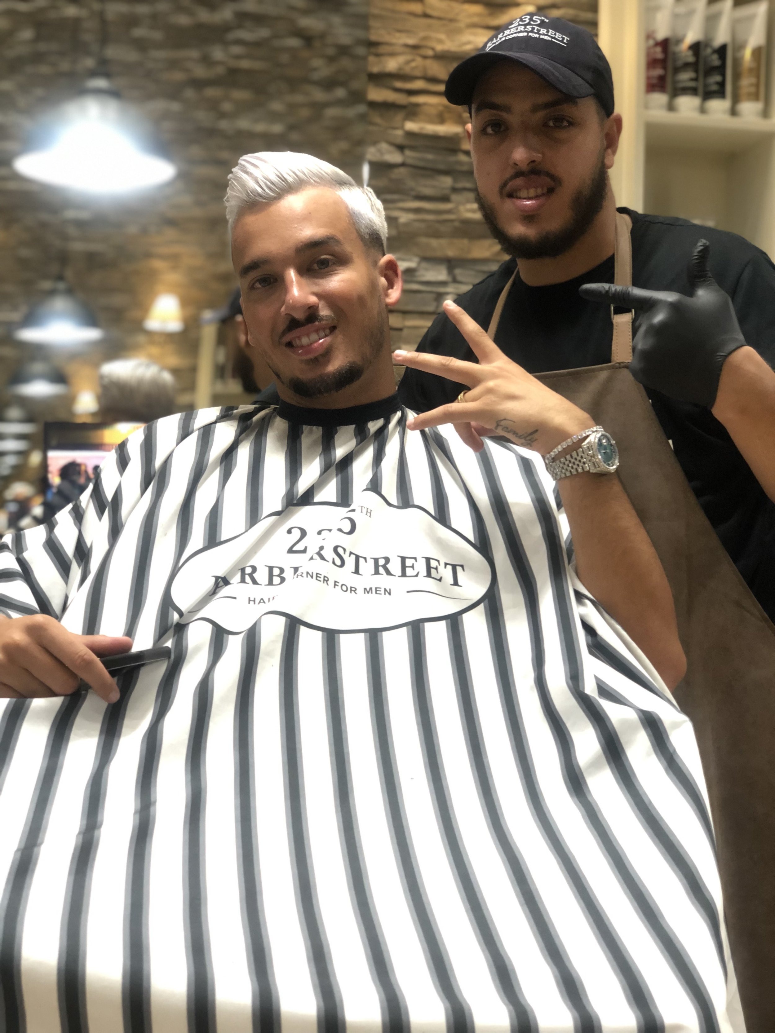 Un homme avec des cheveux gris et une barbe sourit, assis dans un salon de coiffure avec une cape à rayures blanches et noires. Un coiffeur derrière lui, portant un casque noir, une veste en plastique beige, des gants en latex noirs, fait un geste avec la main. Le fond montre un mur en pierre et des étagères avec des produits pour cheveux.
