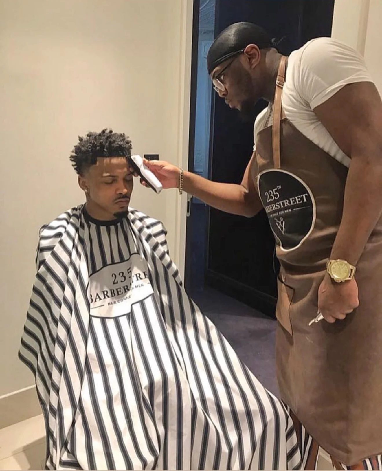 Un homme reçoit une coupe de cheveux dans un salon de coiffure. Il porte une cape rayée et un signe indiquant '235 Barbershop'. Best barber shop salon de coiffure Marrakech