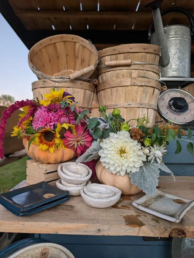 Arrangement of colorful flowers in pumpkins, surrounded by stacked wooden baskets and kitchen scale on a wooden surface.