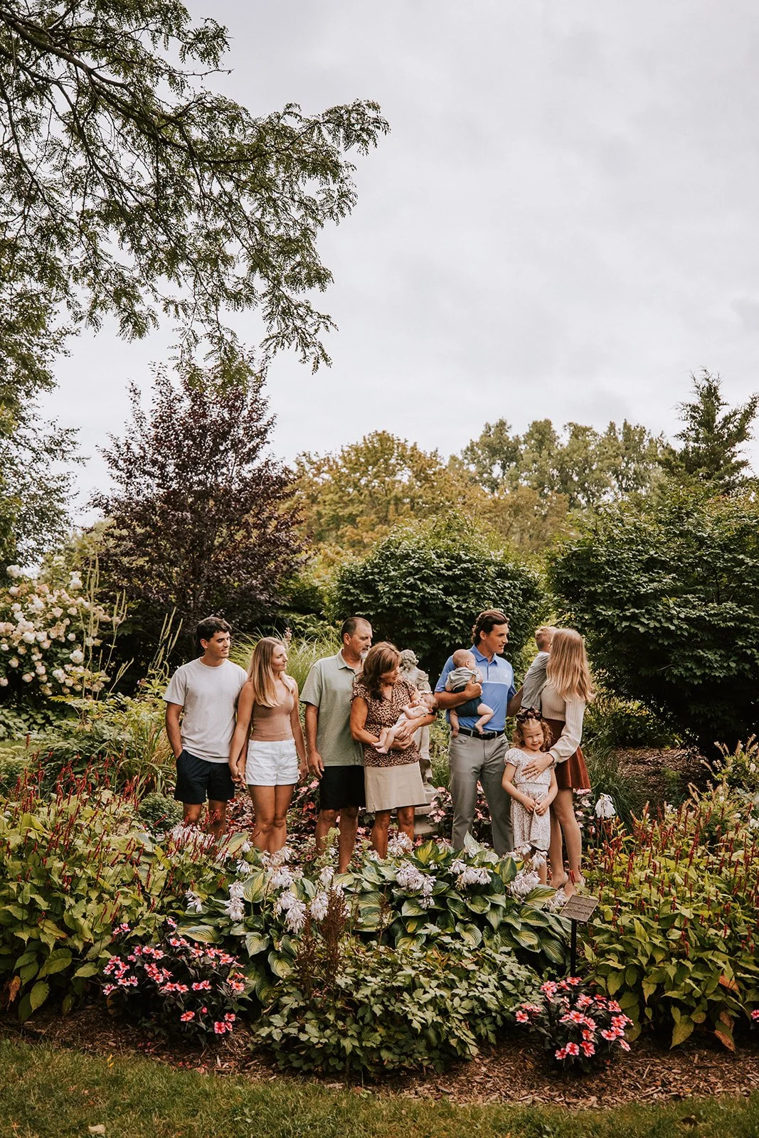A family of diverse ages and genders standing together in a lush garden with blooming flowers and green trees. The sky is overcast.