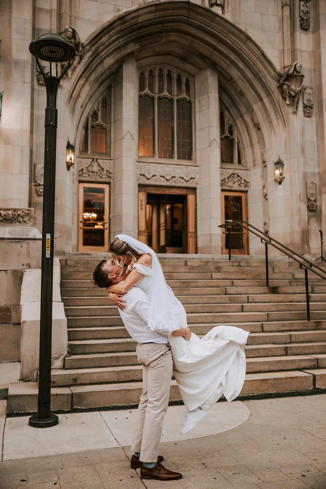 A joyful couple on their wedding day, with the groom lifting the bride in his arms in front of a historic stone building with stairs and gothic windows.