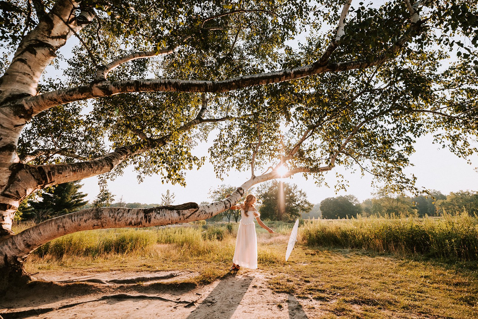 A woman in a white dress holding a white parasol under a large tree on a sunny day, with sunbeam shining through the leaves