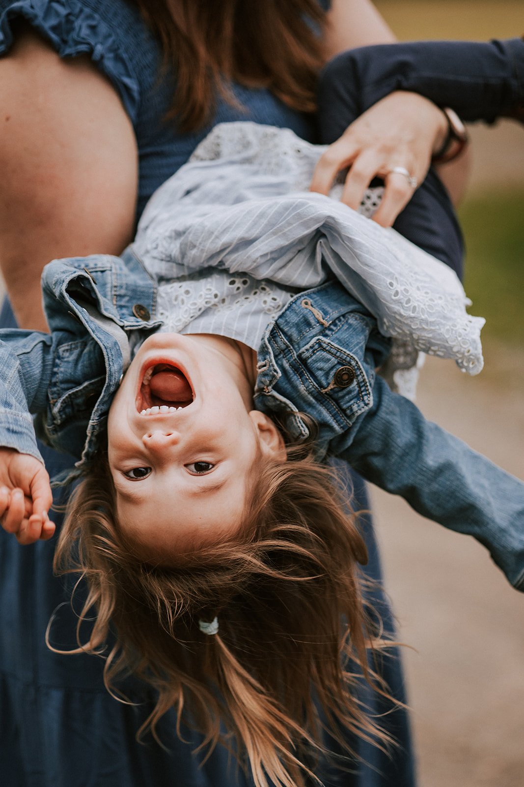 A young girl with brown hair is being held upside down by an adult, smiling and showing her teeth, while the adult's face is not visible in the image.