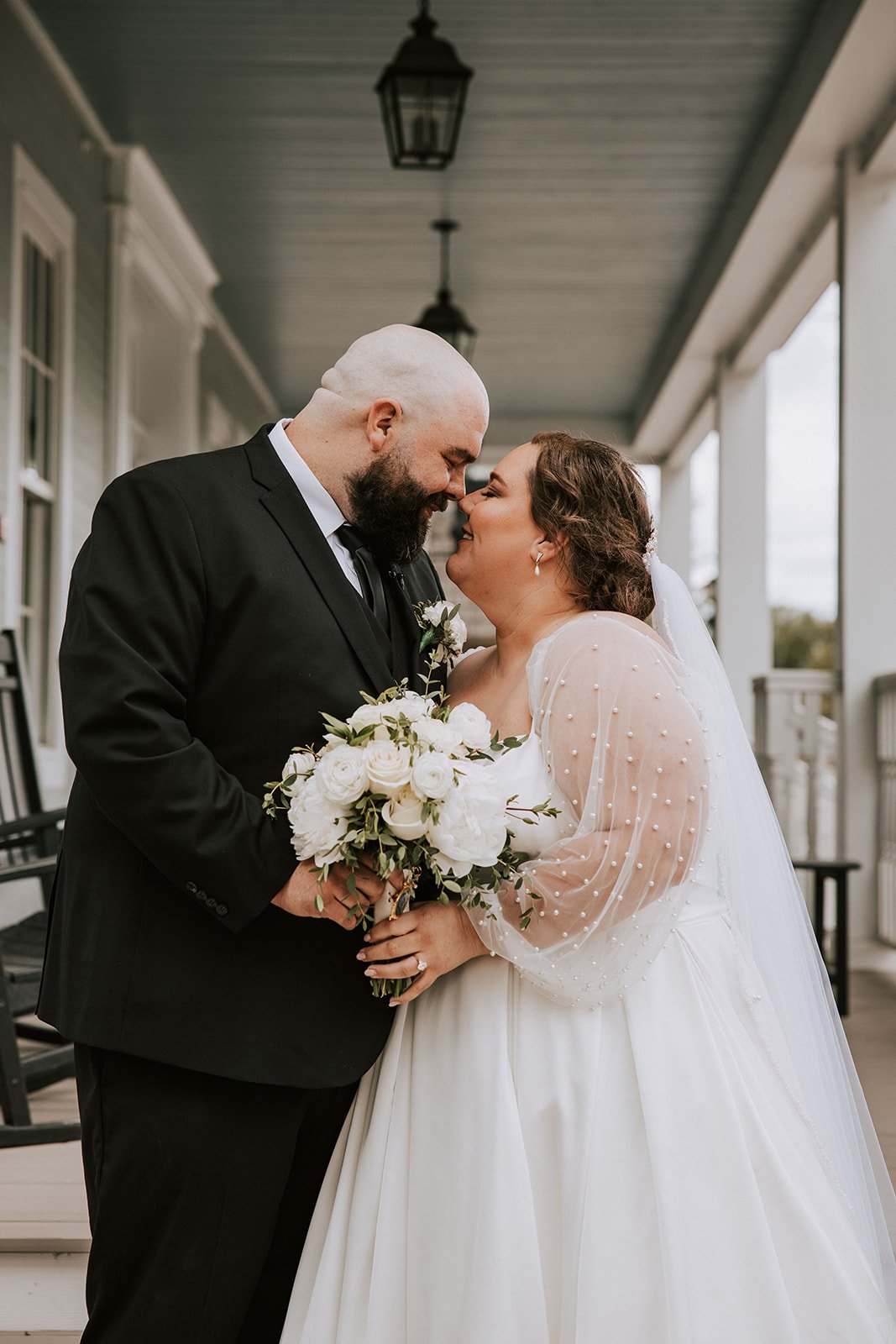 A bride and groom are standing close together on a porch, touching their foreheads and nose, holding a bouquet of white roses and greenery, and smiling at each other during their wedding.
