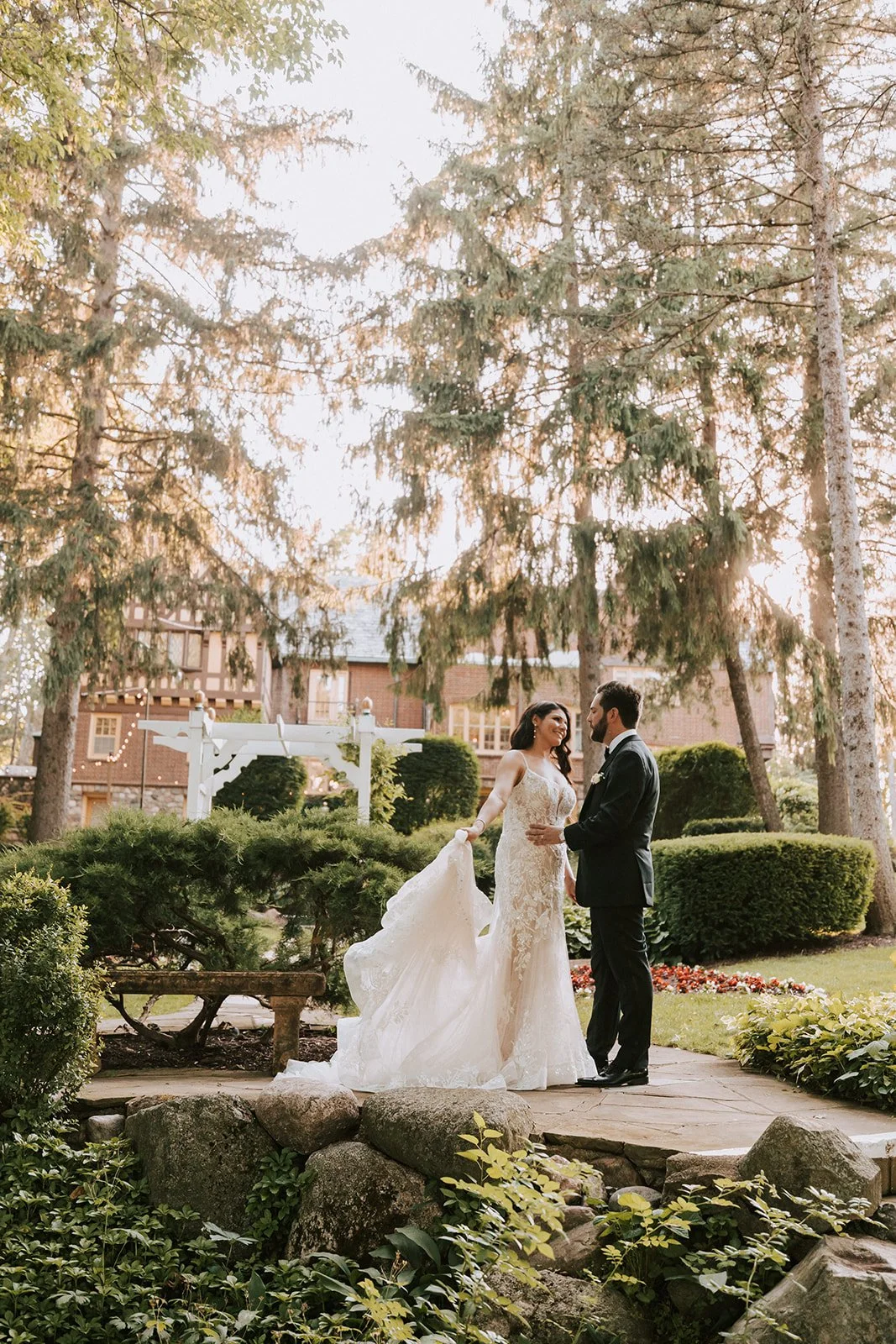 A bride and groom standing close together outdoors in a garden, surrounded by trees and greenery, during sunset.