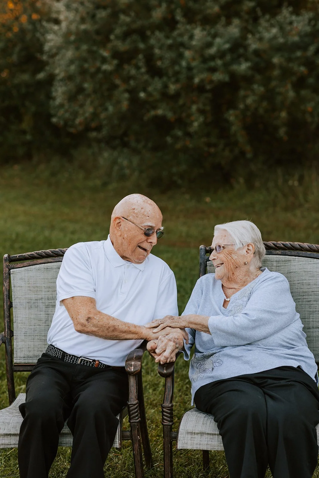 An elderly man and woman sitting outdoors on lawn chairs, smiling and holding hands, sharing a joyful moment.