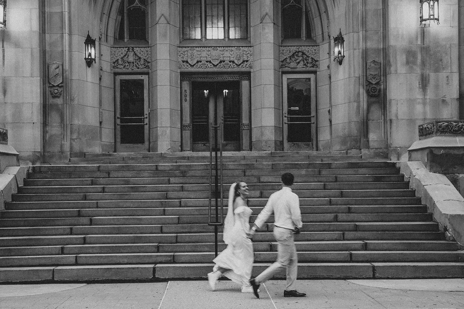 A couple dressed in wedding attire holding hands and walking down the steps of a historic building, possibly a church or courthouse, with ornate stonework and large doors in the background.