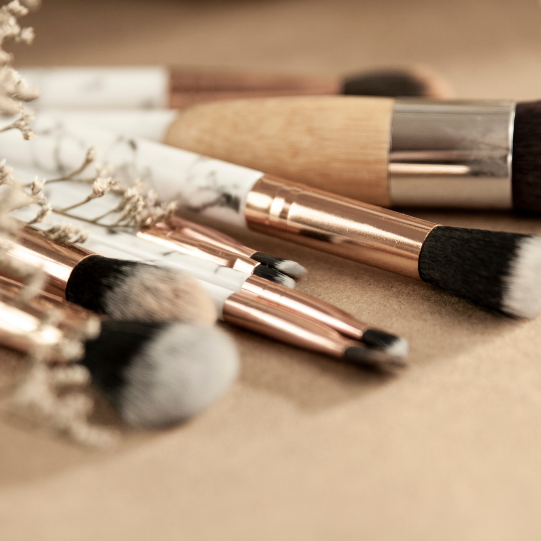 Collection of makeup brushes with rose gold accents, lying on a brown surface, with some dried flowers nearby.