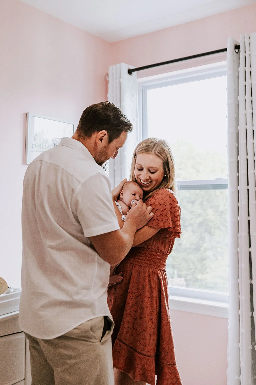 A happy family with a man, woman, and baby standing near a window in a bright room.
