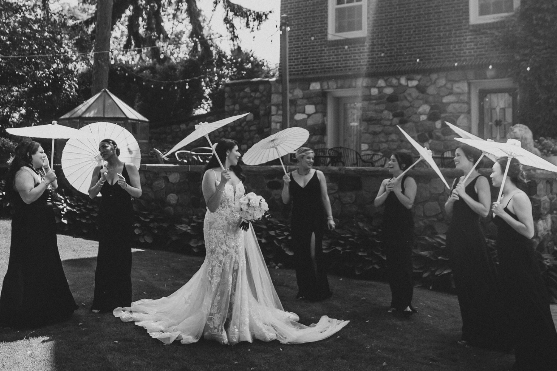 Bride holding a bouquet and wearing a wedding dress, standing outdoors with six bridesmaids dressed in black, holding umbrellas and standing in girl group formation.
