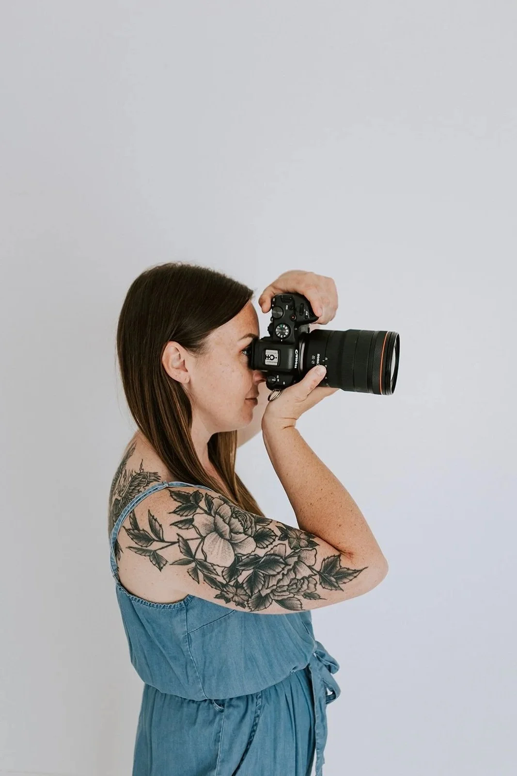 A woman with long brown hair and a floral tattoo on her arm, wearing a blue sleeveless dress, is taking a photograph with a professional camera.
