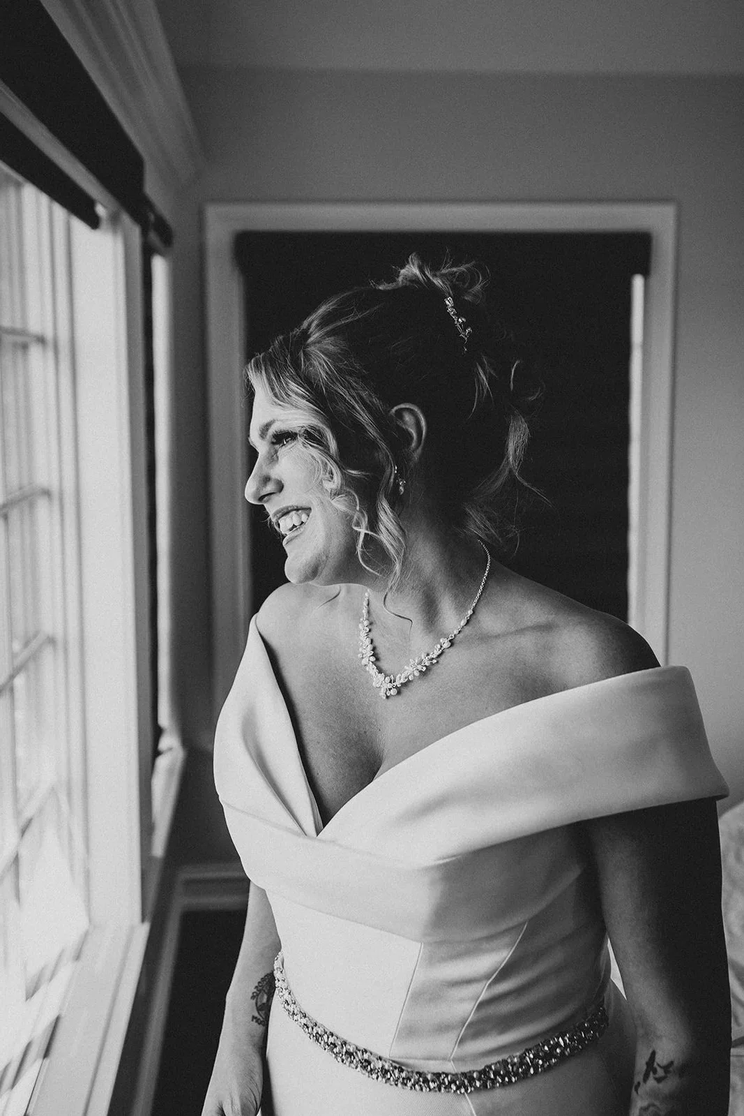 A smiling woman wearing a wedding dress with off-shoulder design and a beaded waistline, looking out a window in black and white.