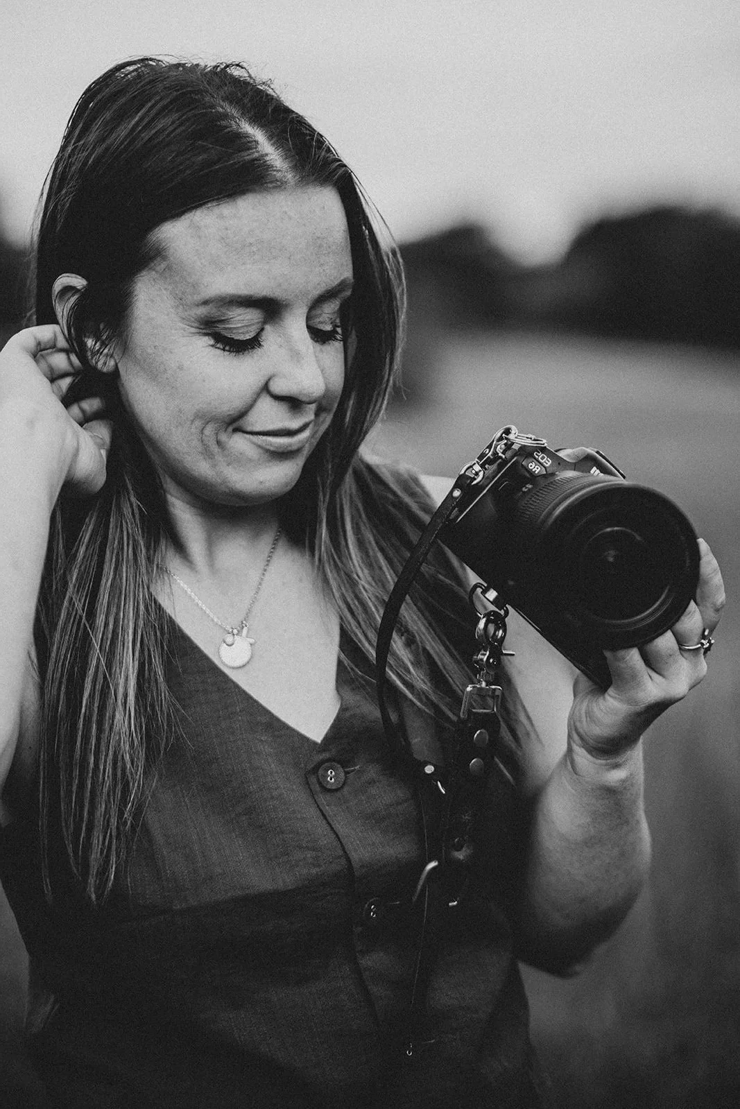 A woman with long hair is holding a camera and smiling while looking down at it outdoors.