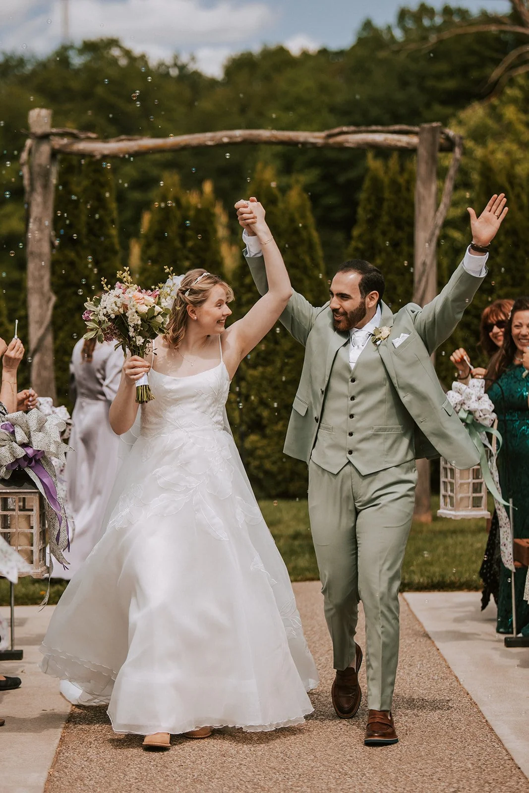 A newlywed couple celebrating as they walk hand-in-hand down the aisle at their outdoor wedding, with the bride holding a bouquet and both smiling joyfully.