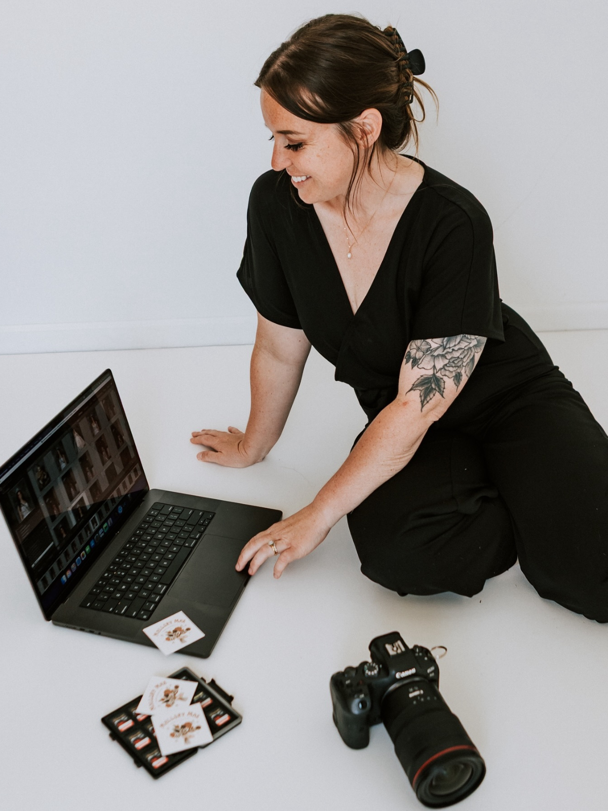 A woman with tattoos on her arm, dressed in black, kneeling on the floor, working on a laptop with photography and editing equipment around her, including a camera and printed photos.