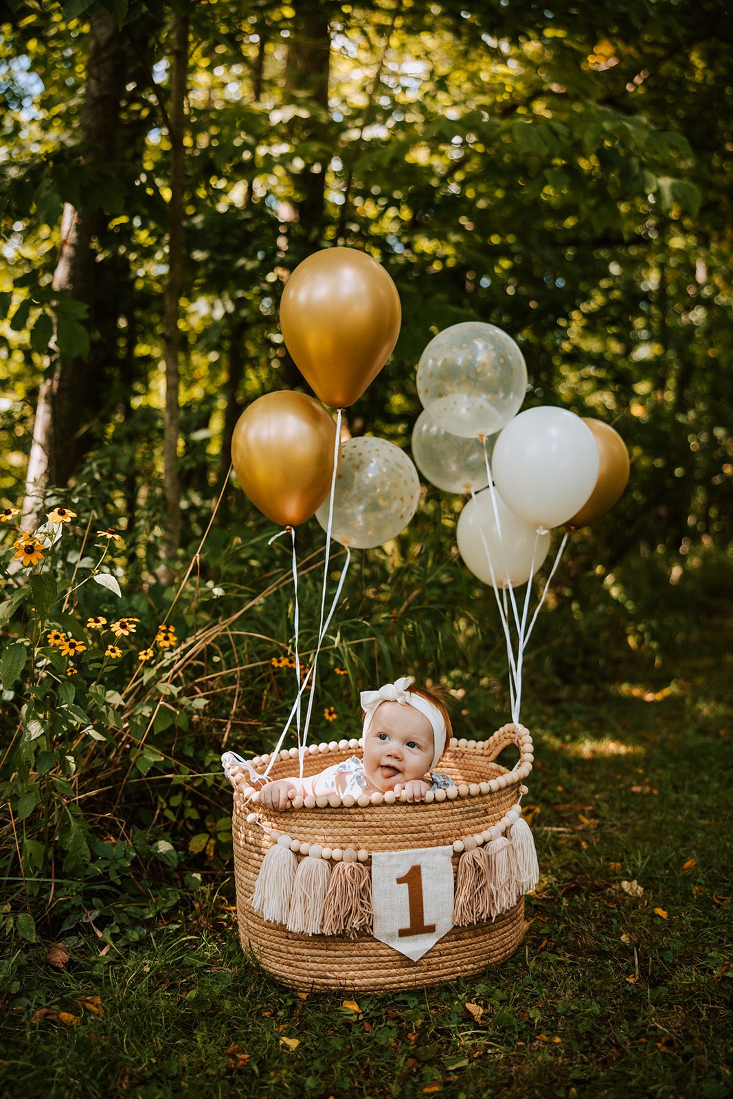Baby girl lying in a woven basket decorated with tassels and a '1' banner, surrounded by gold and clear balloons outdoors in a forest setting.