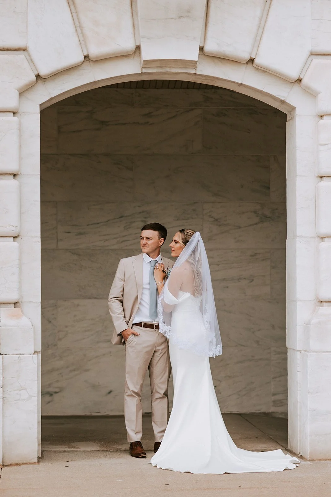 A bride and groom standing under an archway, dressed in wedding attire, with the bride wearing a white gown and veil and the groom in a light-colored suit.