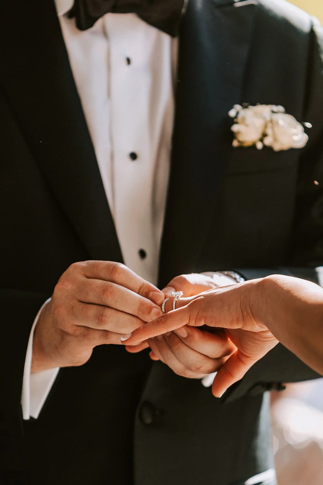 A groom in a tuxedo is placing a wedding ring on a bride's finger during a wedding ceremony.