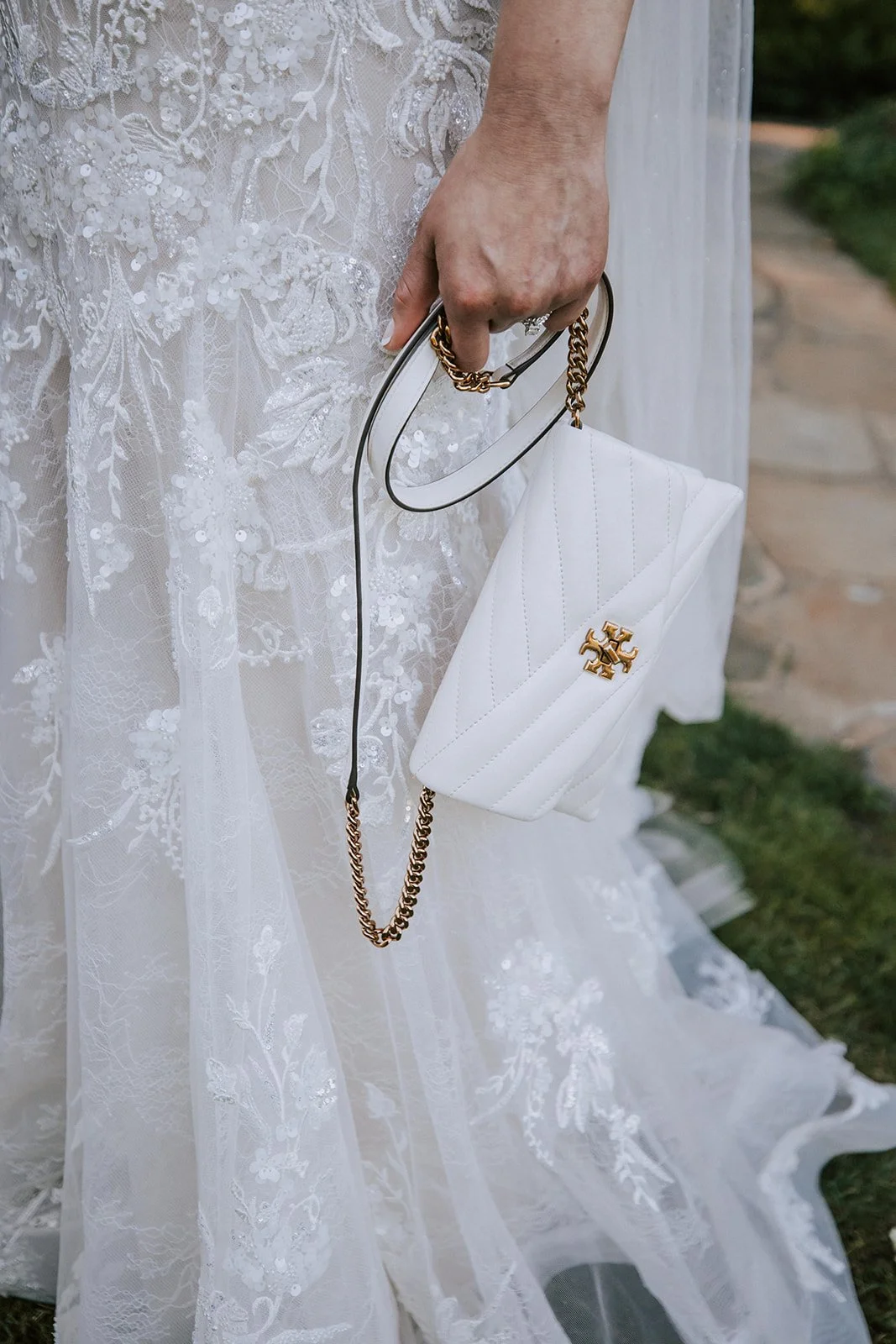 Close-up of a woman wearing a white lace wedding dress, holding a small white quilted purse with a gold logo and a gold chain strap.