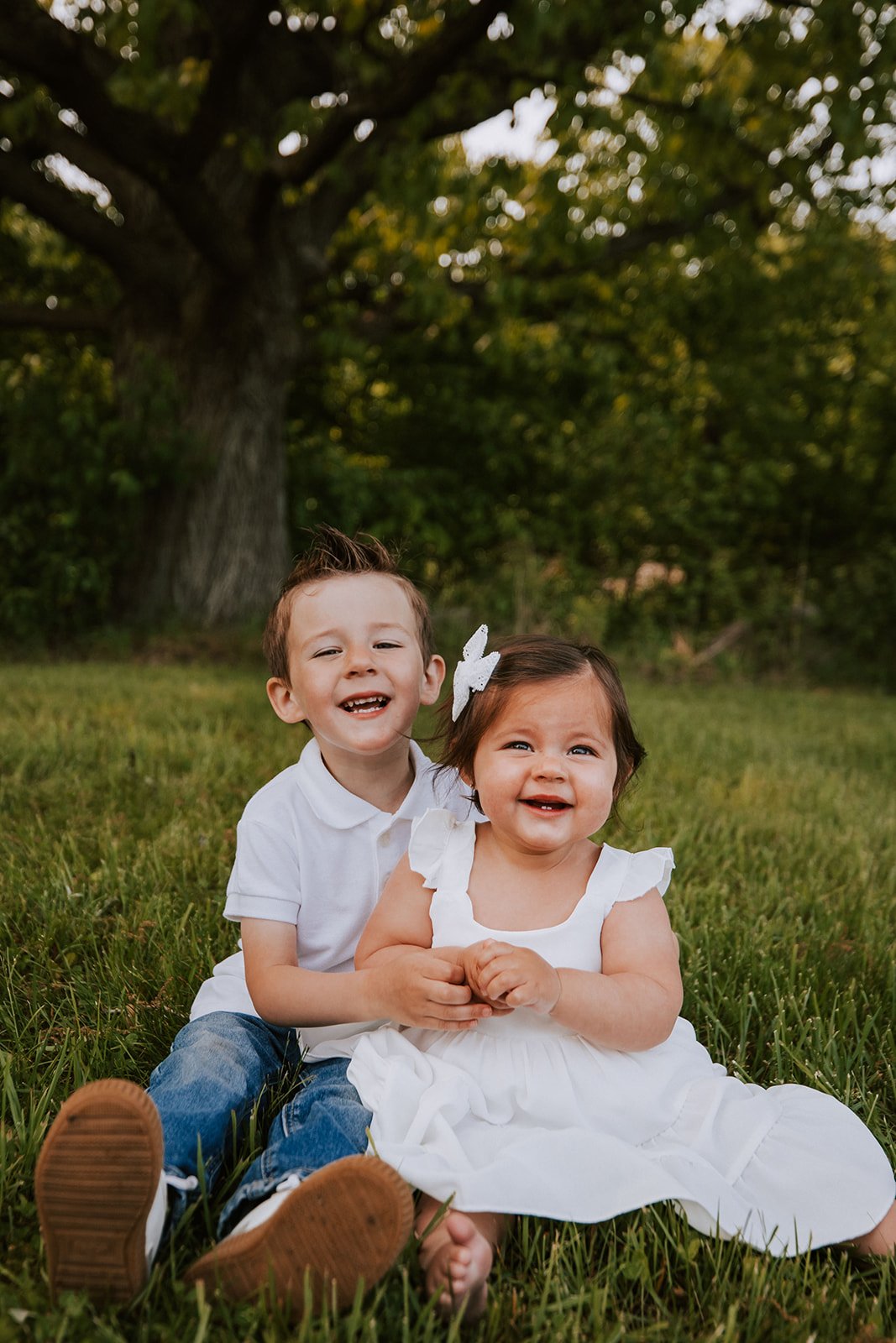 Two young children, a boy and a girl, sitting on grass under a large tree, smiling and holding hands, dressed in white clothing.