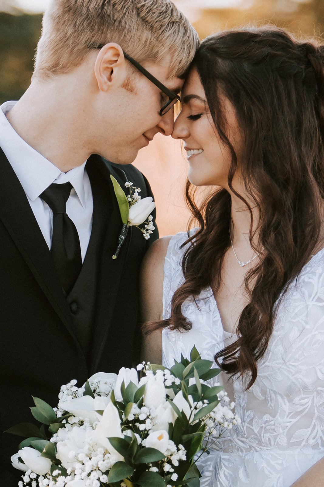 A newlywed couple faces each other with foreheads touching, smiling, with the bride holding a bouquet of white flowers, outdoors during sunset.