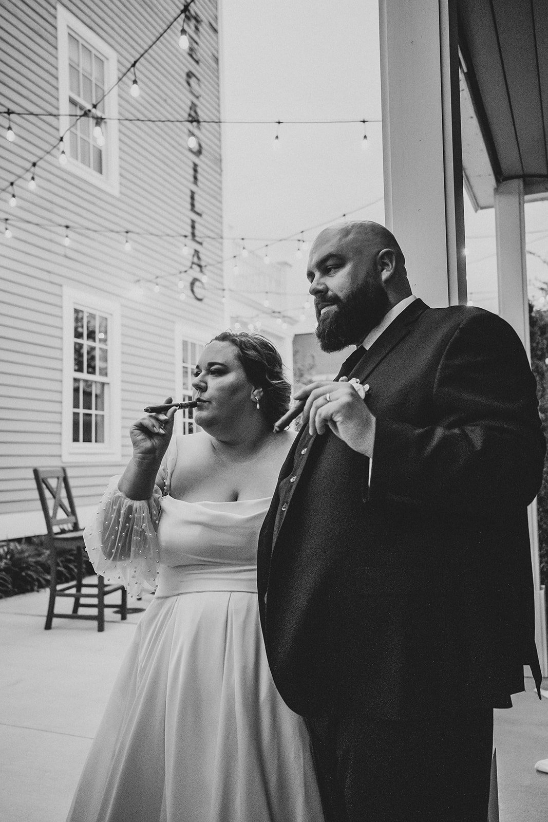 Black and white photo of a woman and a man standing outside, both holding cigars. The woman is wearing a white dress with sheer, polka-dotted sleeves, and the man is wearing a dark suit. Decorative string lights hang above them, and a building with a sign that reads 'COCKTAIL' is visible in the background.