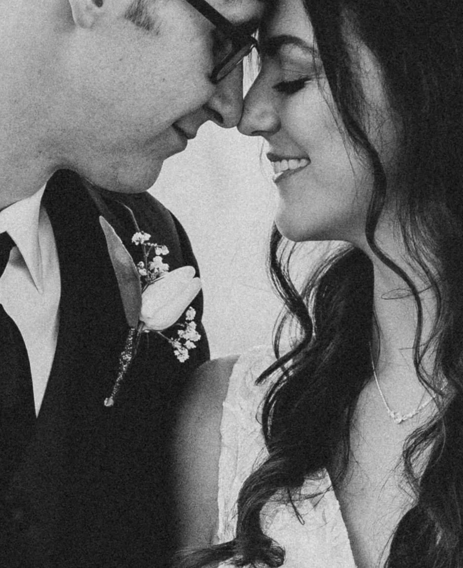 Black and white photo of a couple with the man wearing glasses, a suit with a boutonniere, and the woman with long wavy hair smiling.