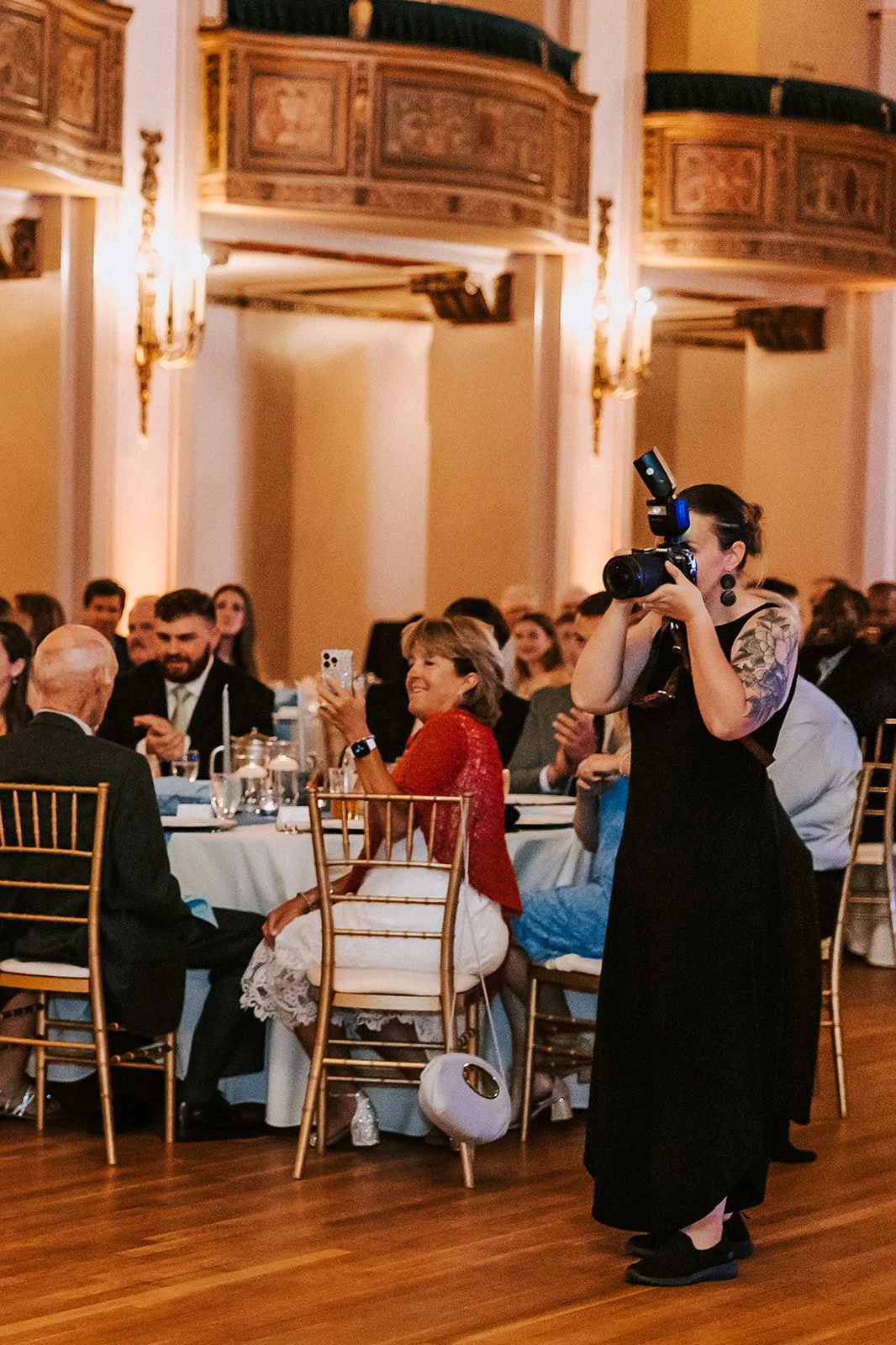 A woman with tattoos on her arm taking a photograph at a formal event in a grand ballroom with gold chairs and chandeliers.