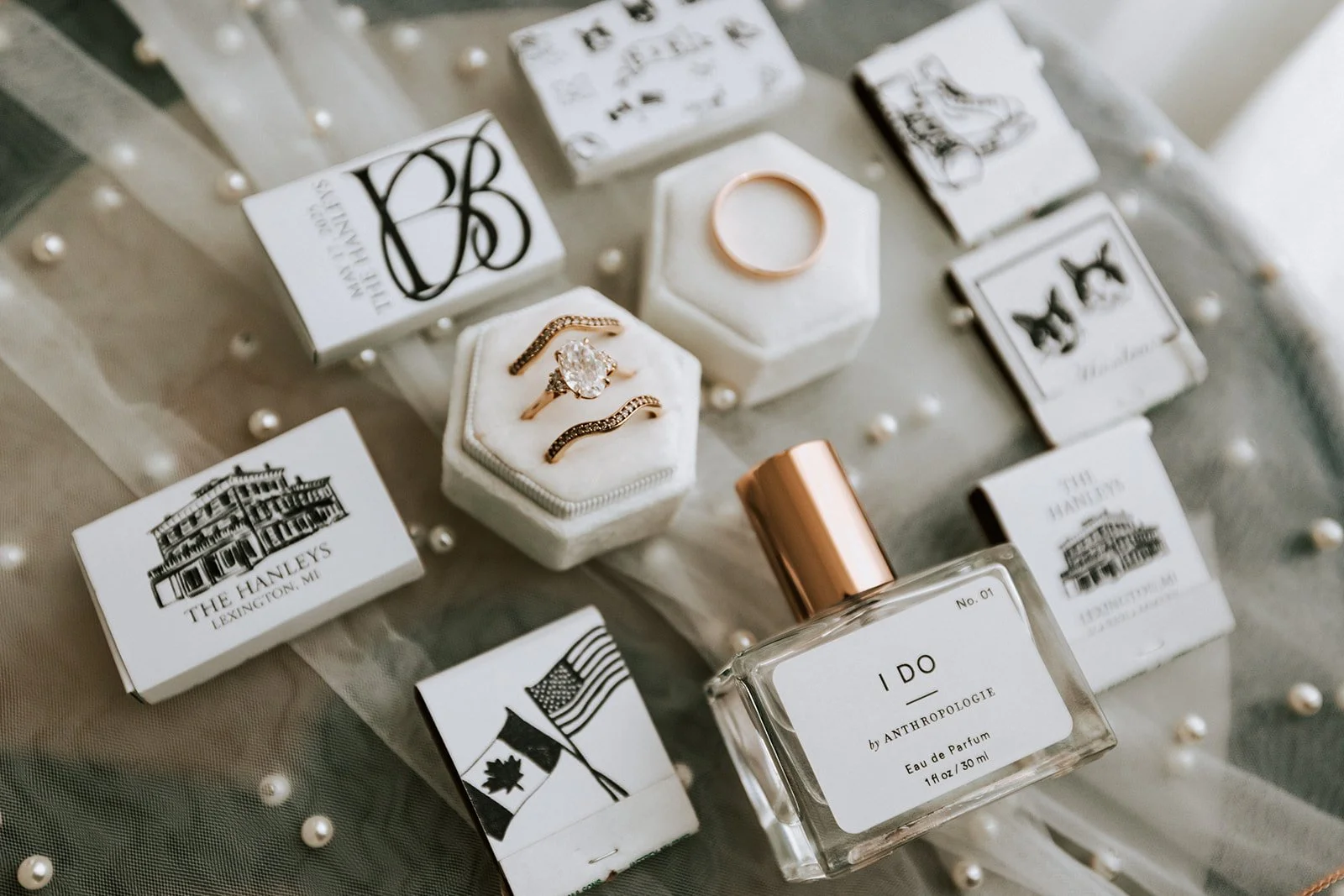 Collection of jewelry, perfumes, and decorative boxes on a table. The jewelry includes rings and earrings displayed on a white hexagonal pillow. The perfume bottle labeled I DO is prominent, and there are small decorative boxes with black and white illustrations and text.