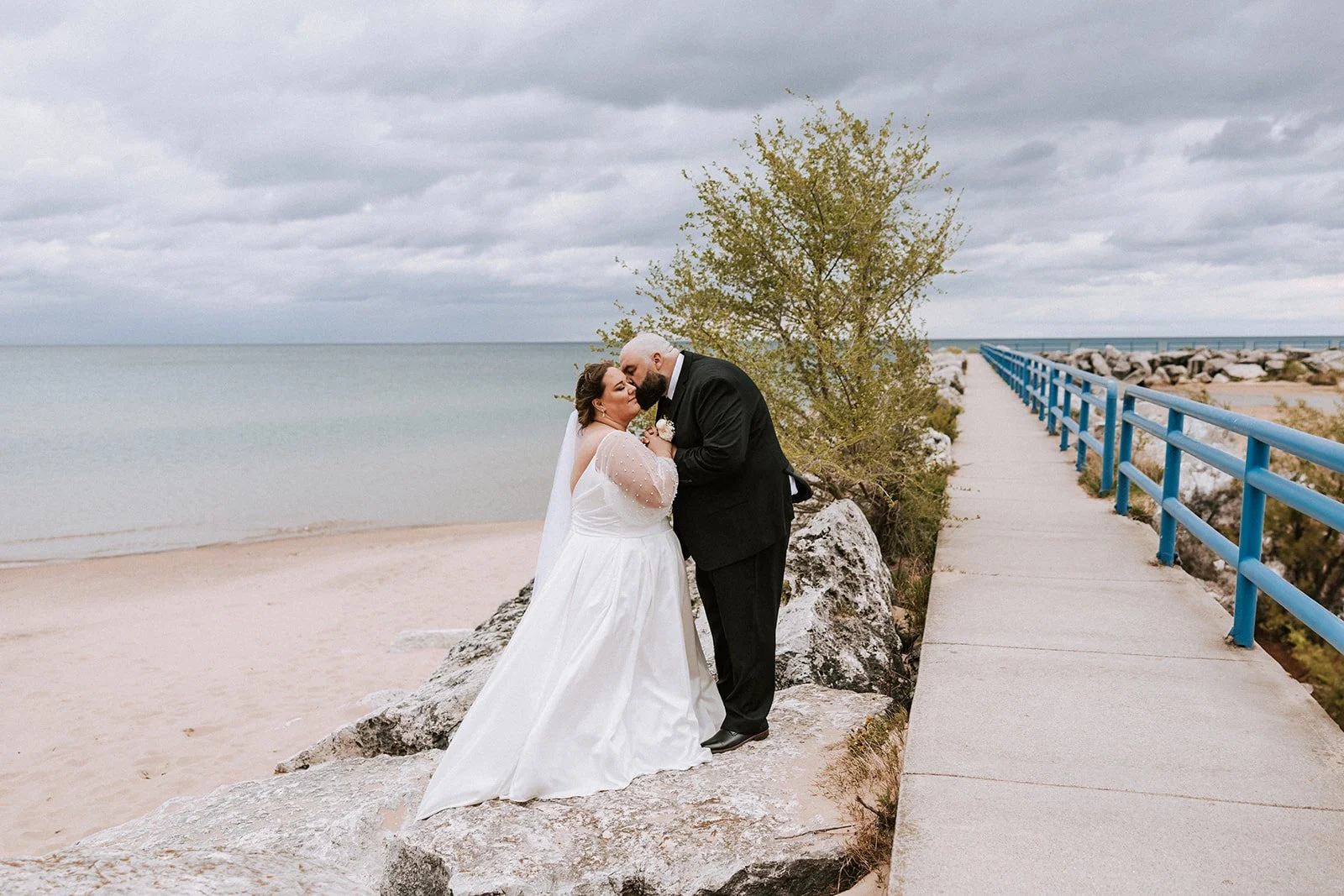 A bride and groom sharing a kiss on a rocky beach with the ocean and cloudy sky in the background, near a blue railing.