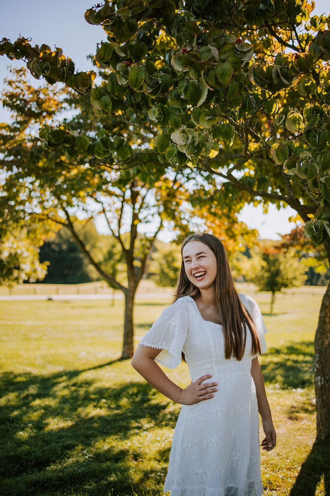 A young woman in a white dress standing outdoors under a tree, smiling and laughing with her hand on her hip in a sunny park with green grass and trees.