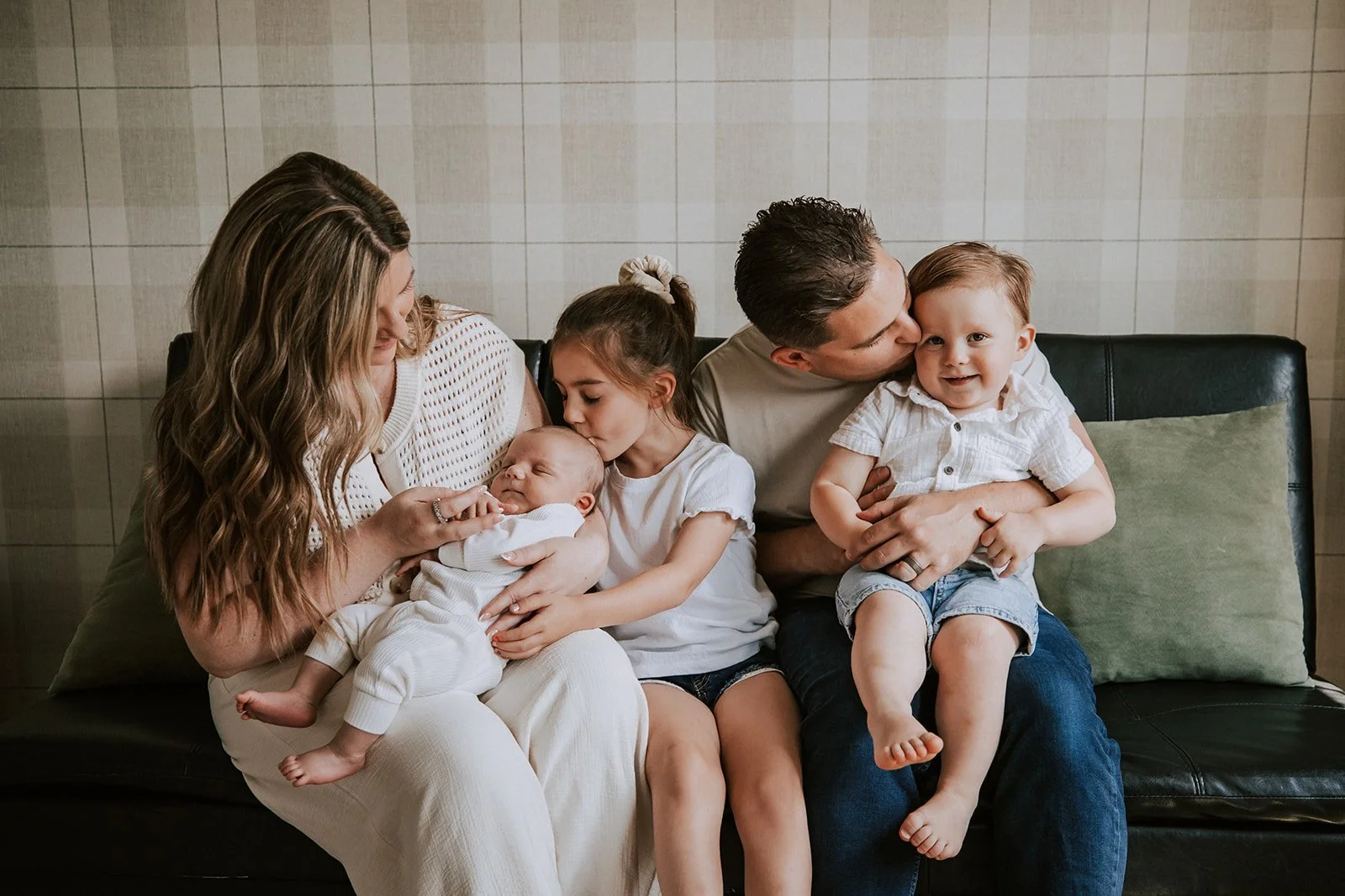 A family of five enjoying a moment on a black leather couch against a beige tiled wall. The mother, with long wavy hair, holds a sleeping baby in white clothing. The older daughter, with her hair in a ponytail, kisses the baby. The father, with short dark hair, kisses a smiling toddler boy on the cheek; the boy wears a short-sleeve shirt and shorts. A green pillow is on the couch.