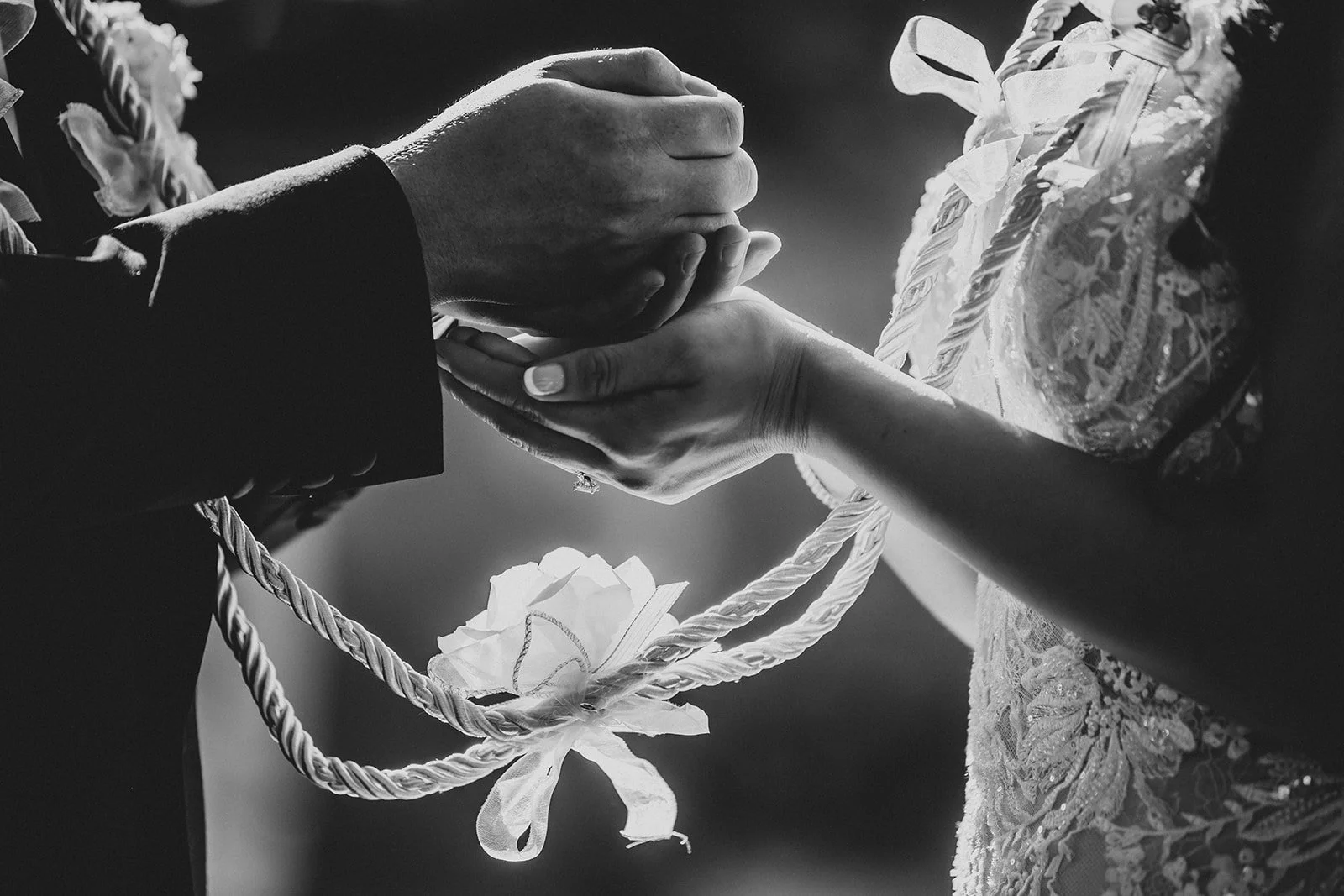 A black and white photo of a wedding ceremony where a man in a suit holds a woman's hand, who is wearing a lace dress with intricate embroidery, and a decorative flower with ribbon is seen in the background.