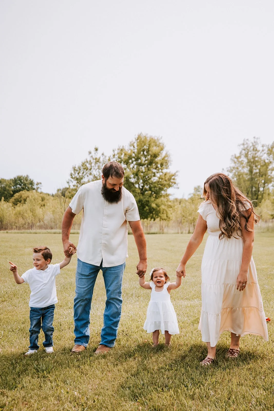 Family of four holding hands and walking in a grassy field, trees in the background, woman in a white dress, man in a white shirt, children in white, sunny day.