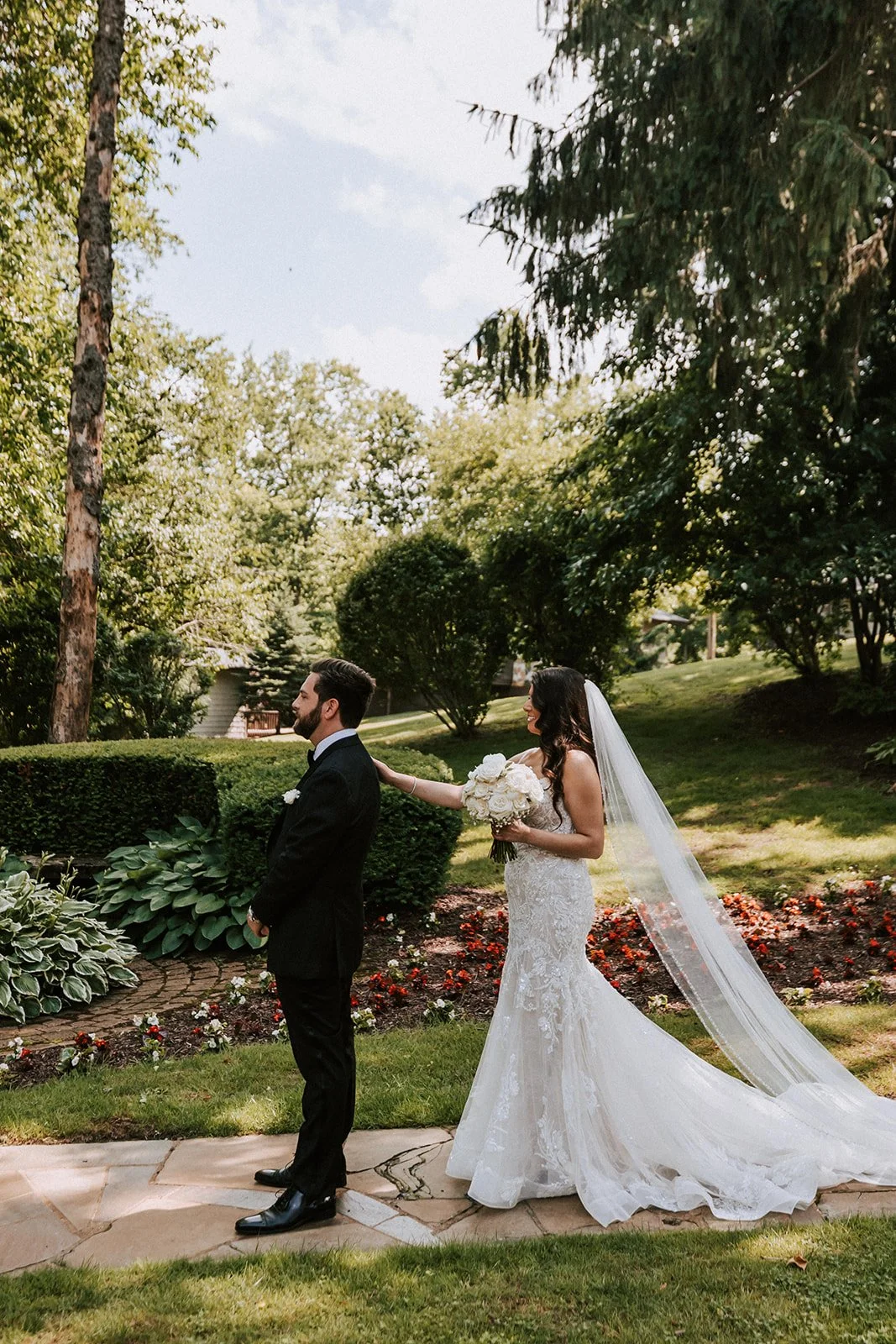 A bride in a white lace wedding dress and veil touched a groom in a black tuxedo with a white boutonniere, standing outdoors in a garden with trees, bushes, and flowers on a sunny day.