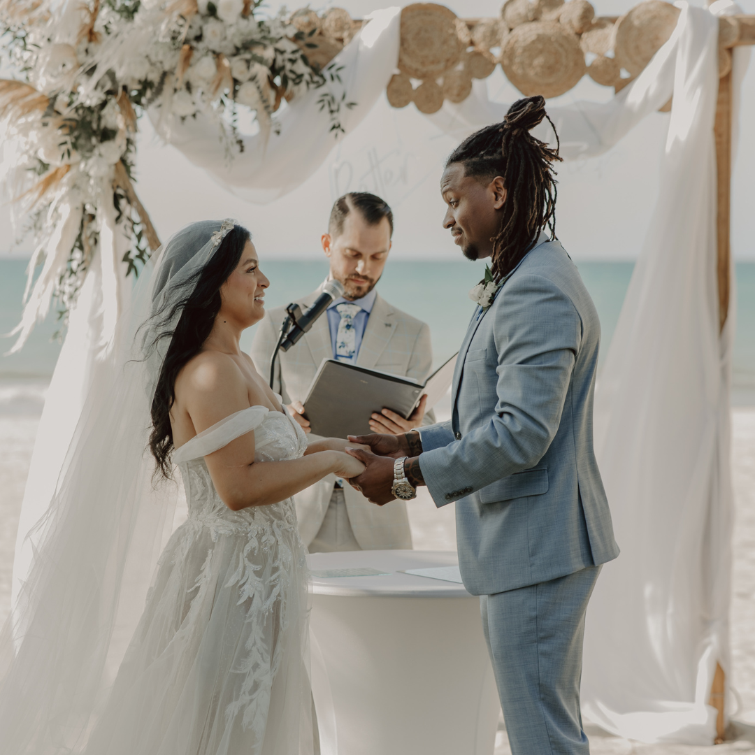 A couple getting married on the beach under a decorated arch, holding hands and facing each other as the officiant reads vows.