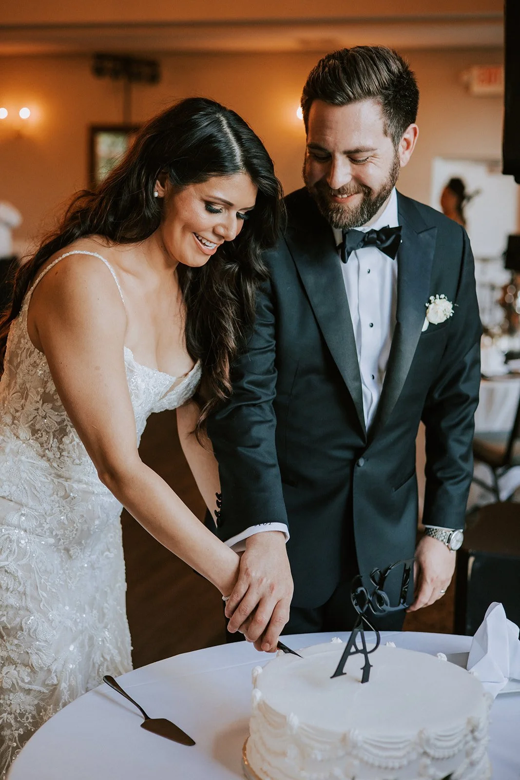 A newlywed couple, the bride in a lace wedding gown and the groom in a tuxedo, smiles as they cut a white wedding cake together.