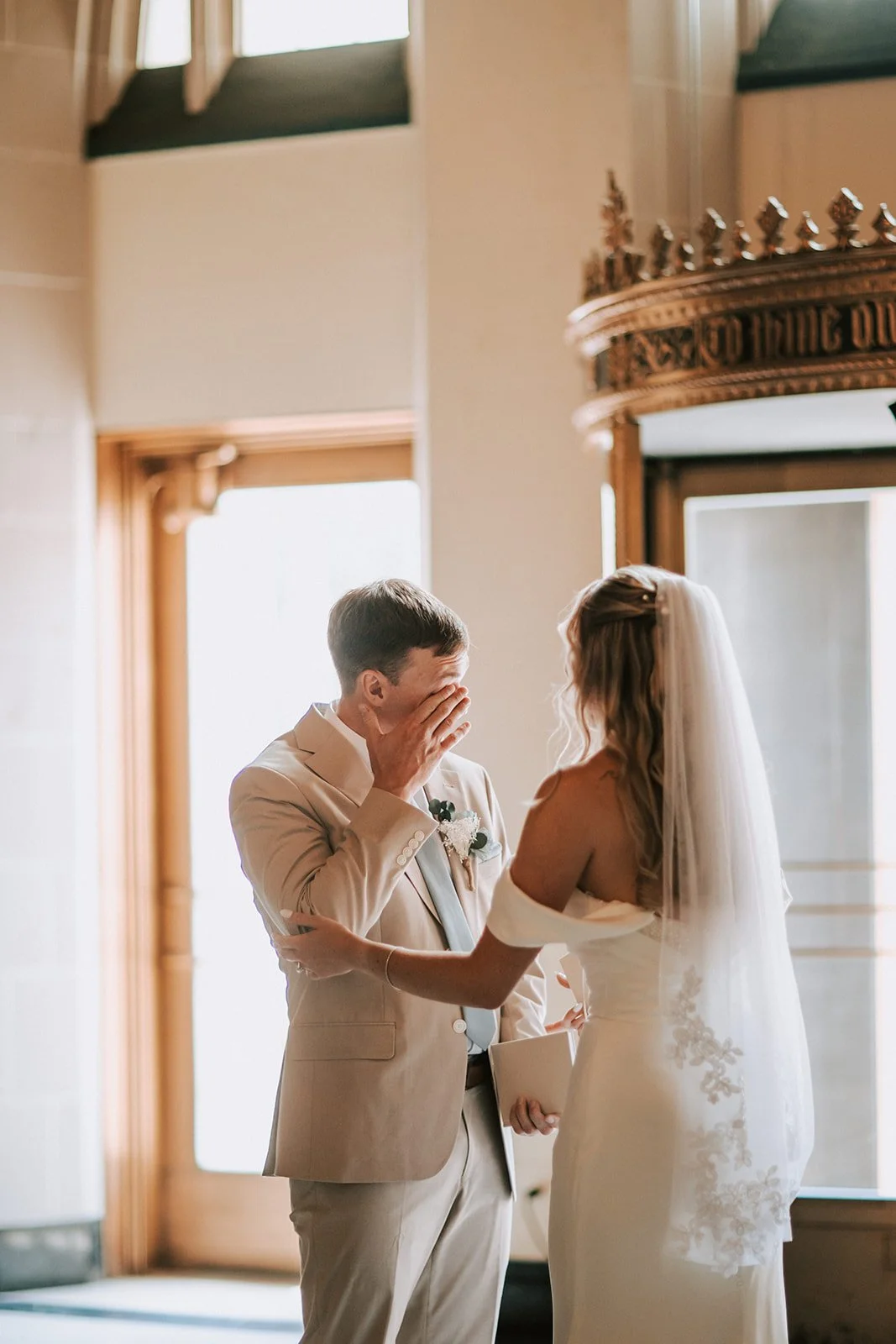 A bride and groom at their wedding ceremony, standing indoors near a window, with the groom holding his face with his hand and the bride touching his arm, both smiling.