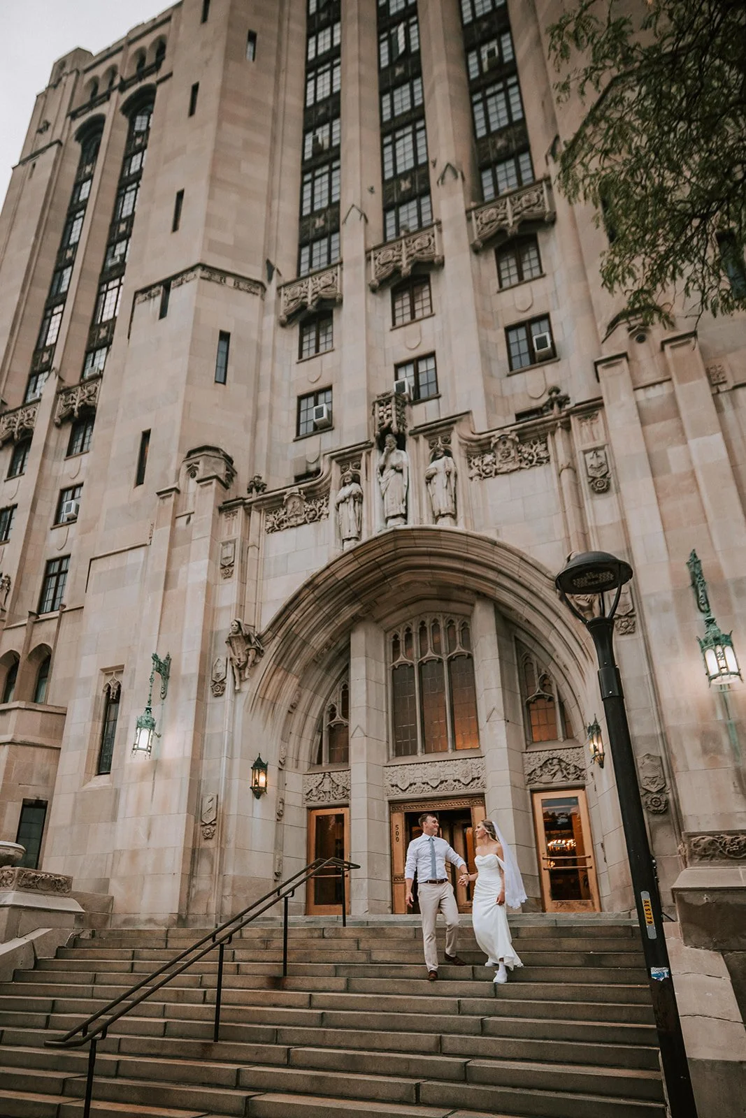 A bride and groom walking down the steps of a historic building with ornate stone carvings and sculptures above the entrance, late in the evening.
