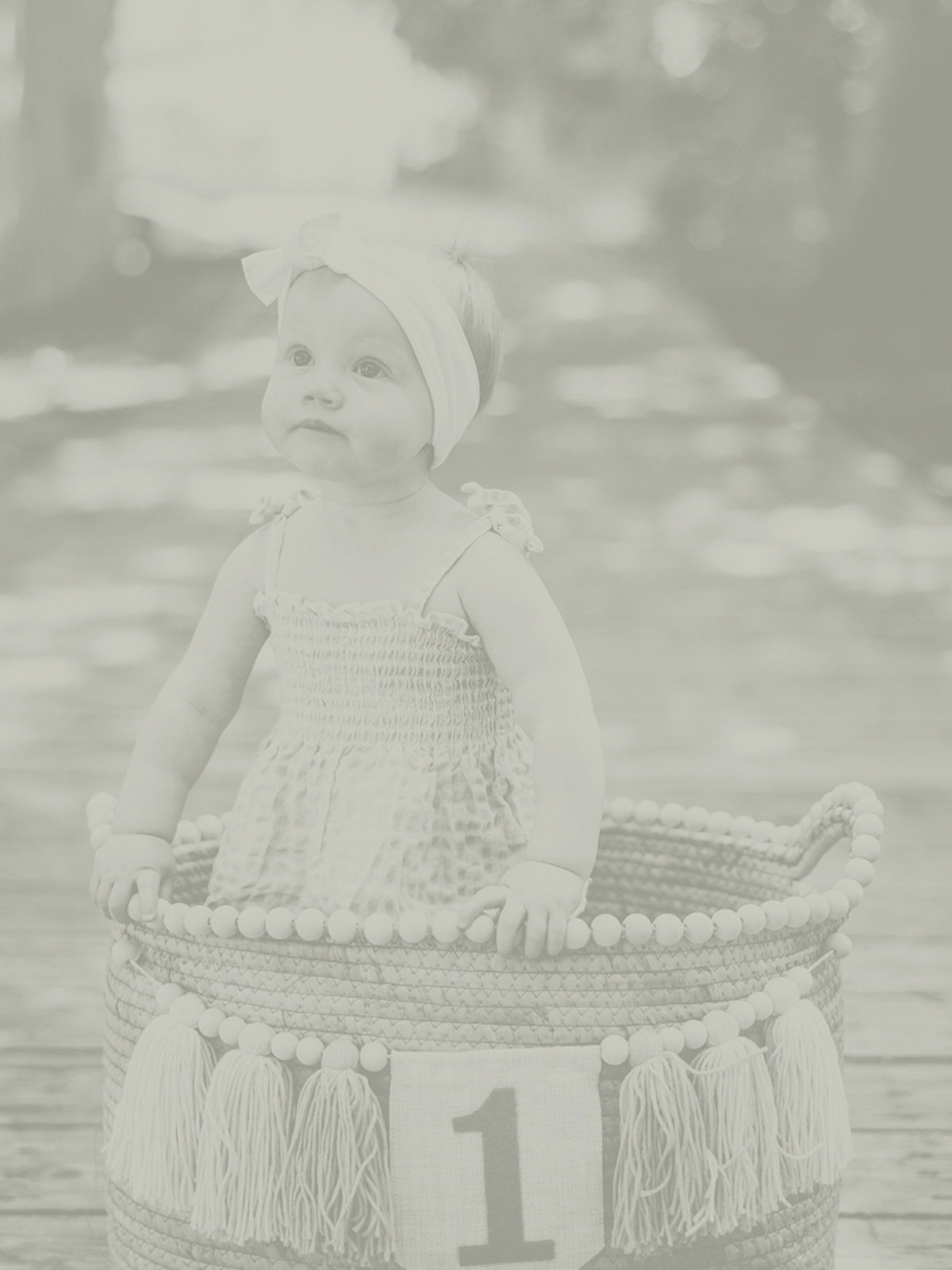 A baby girl sitting in a woven basket with a number 1 on it, outdoors on a wooden deck, wearing a headband and dress, looking curiously to the side.