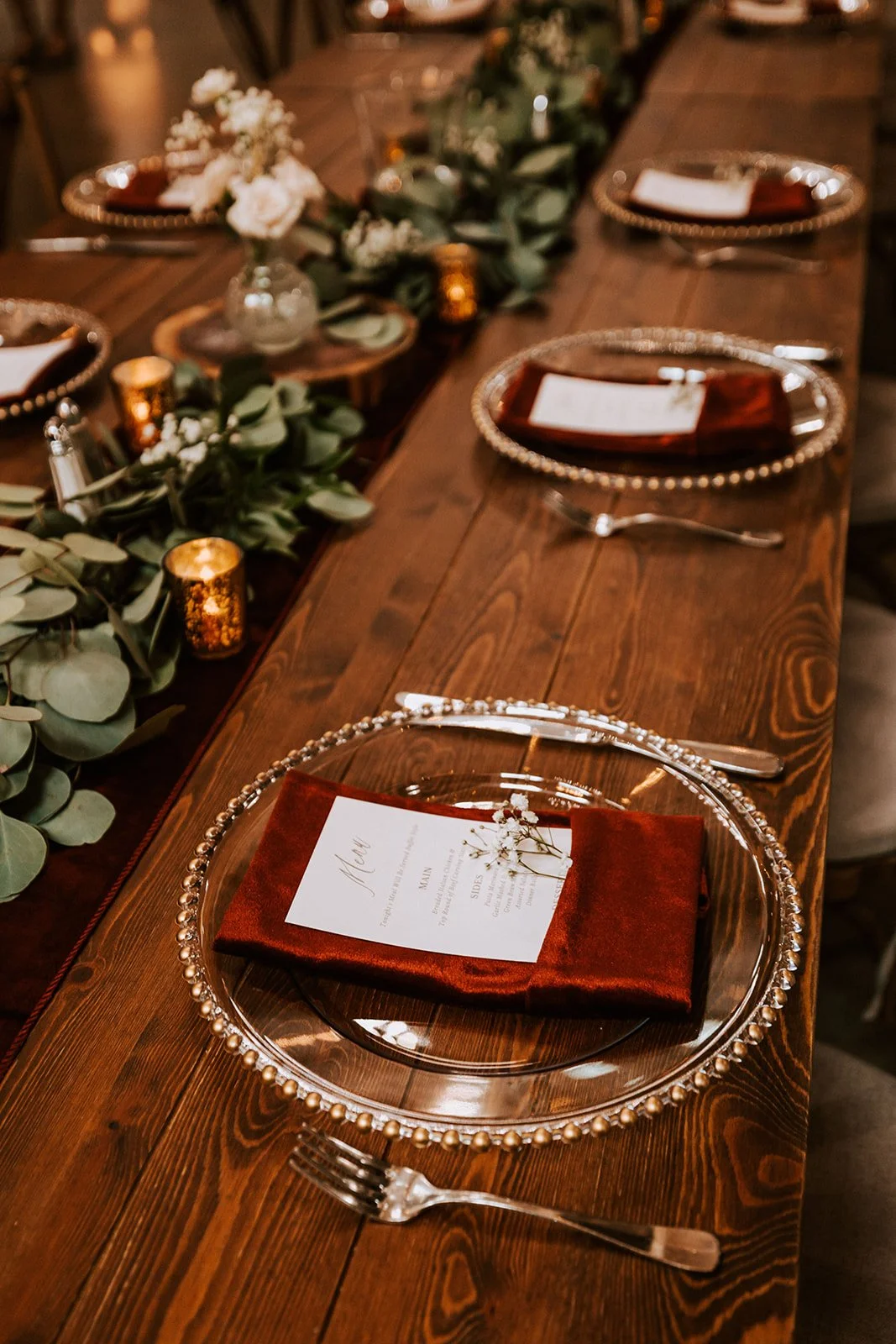A decorated dining table with clear glass plates, red napkins, silver utensils, and a menu on each plate. The table has a green foliage and white flower centerpiece, with small lit candles in gold holders along the length of the table, creating an elegant and warm atmosphere.