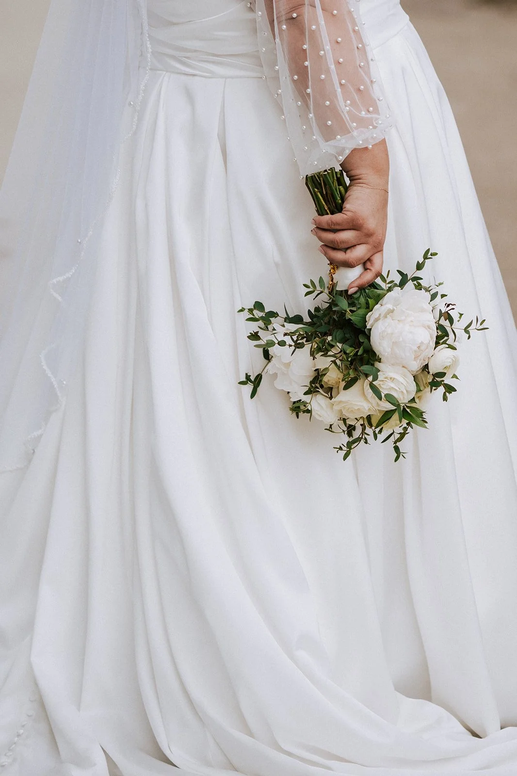 Close-up of a bride holding a bouquet in her right hand, wearing a white wedding dress with a sheer sleeve decorated with pearls.