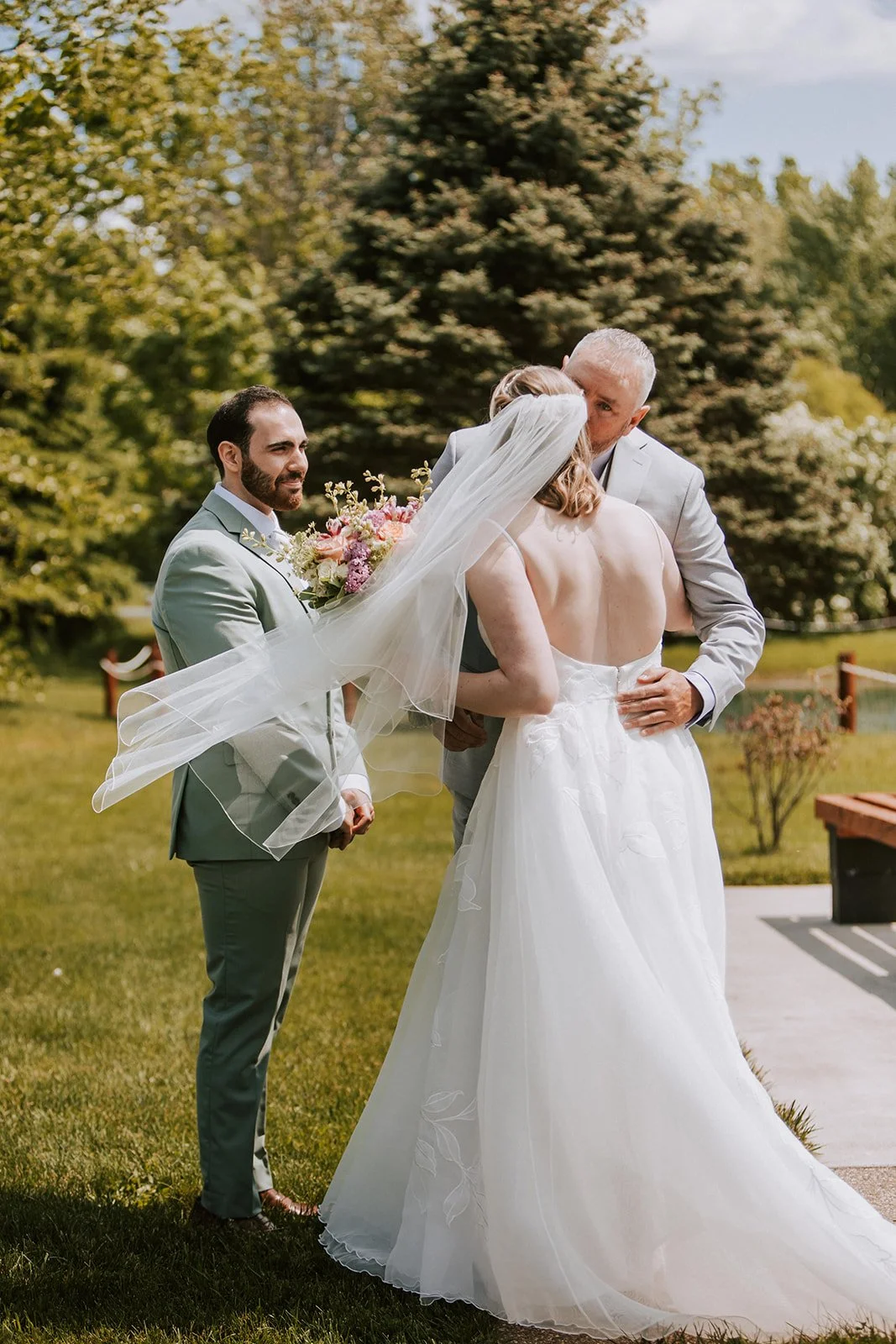 A bride and groom kissing during their wedding ceremony outdoors, with a man holding a bouquet of flowers nearby, all set in a lush green garden.